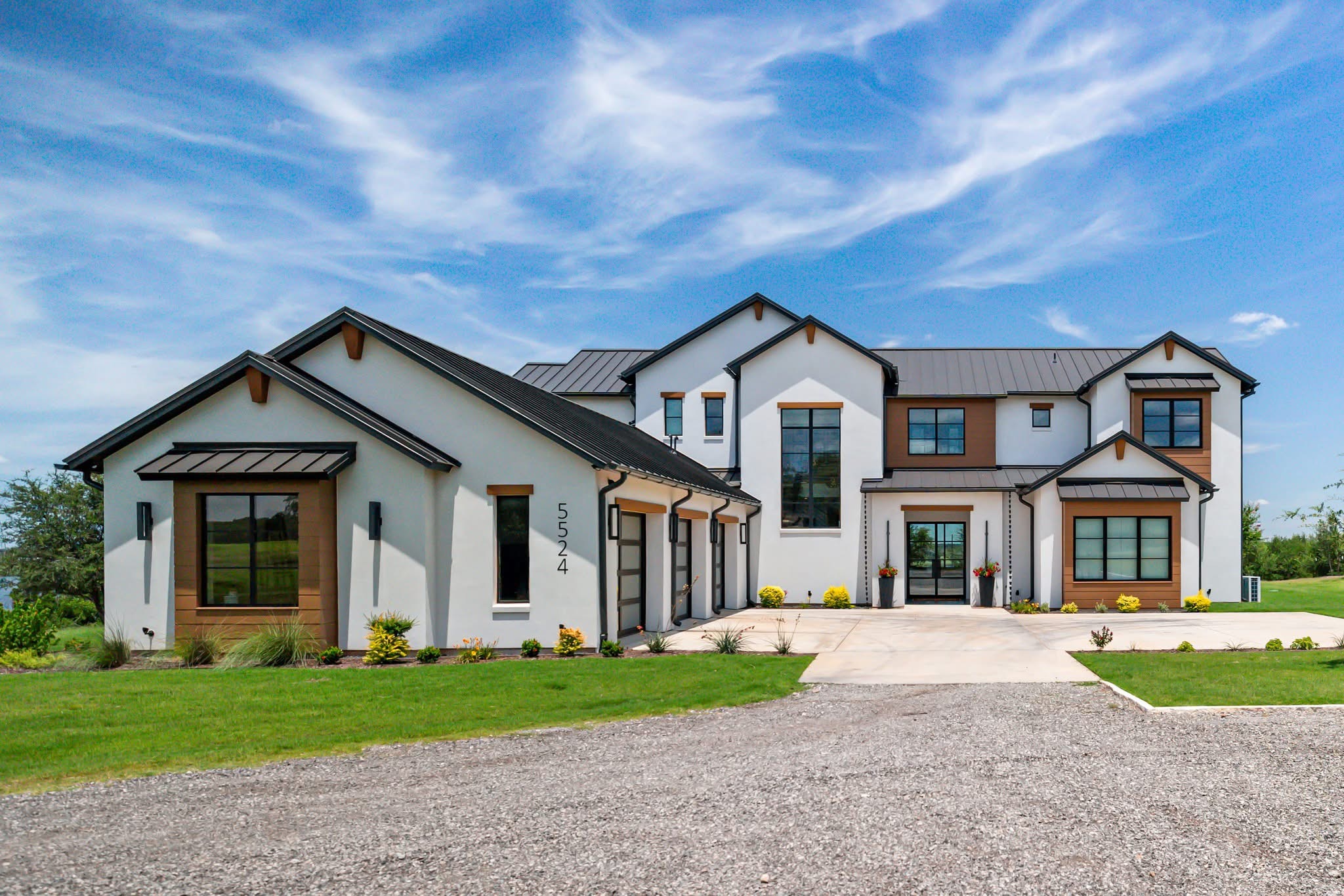 Large modern house with multiple sections, white exterior, brown accents, and a driveway, under a blue sky with clouds.