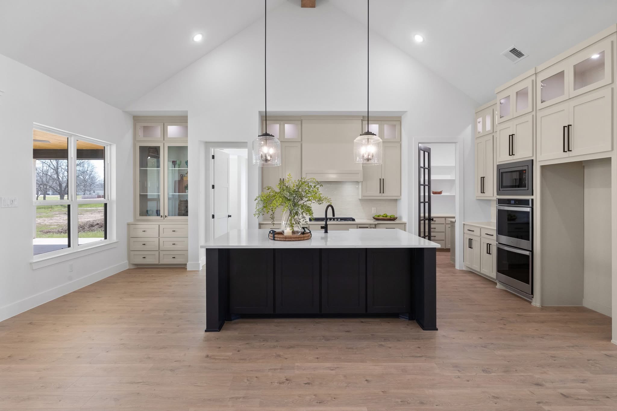 Modern kitchen with white cabinets, black island, pendant lights, and large windows, with light wood flooring and vaulted ceiling.