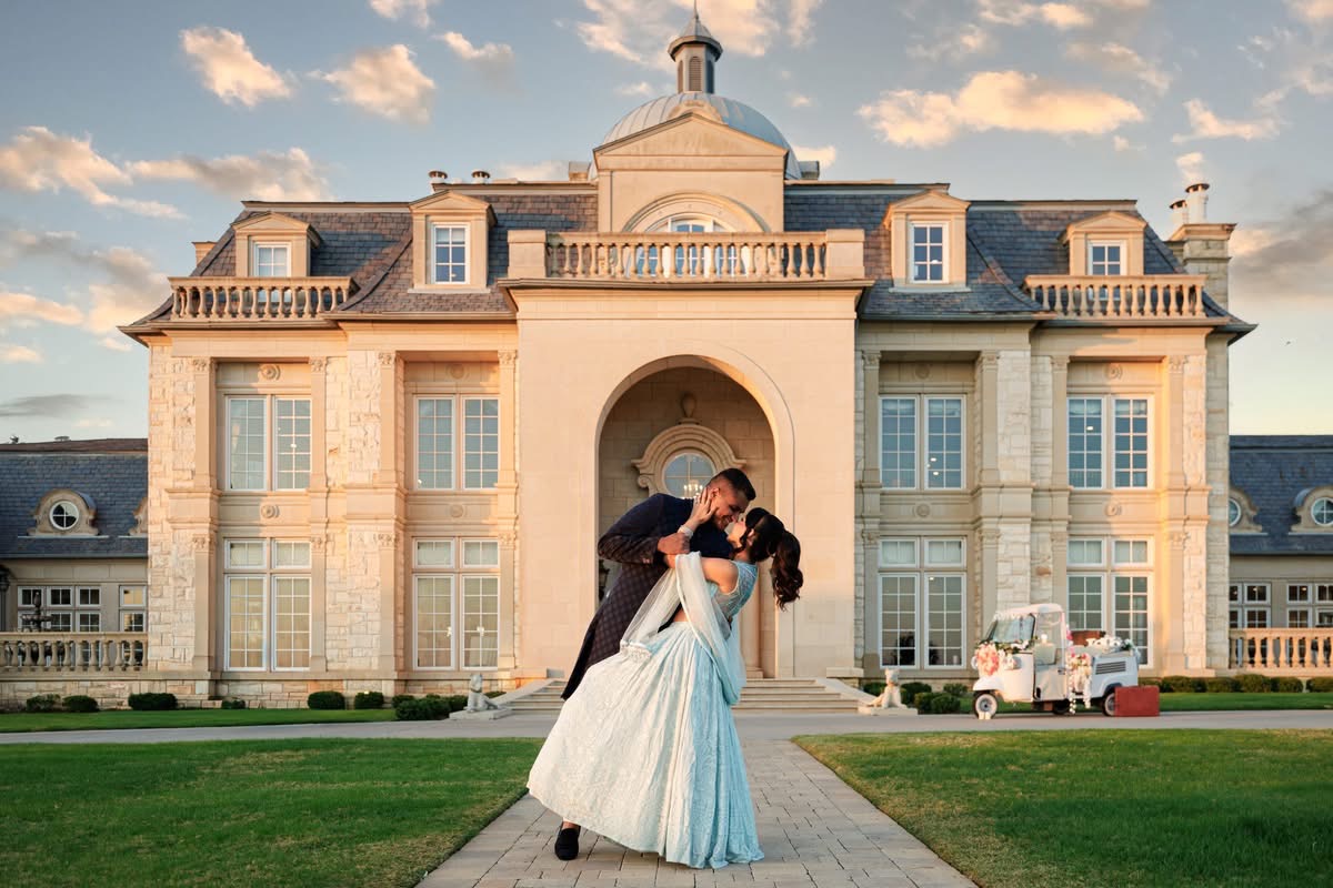 A couple dancing in front of a large, elegant mansion with a central archway and multiple windows, under a partly cloudy sky.