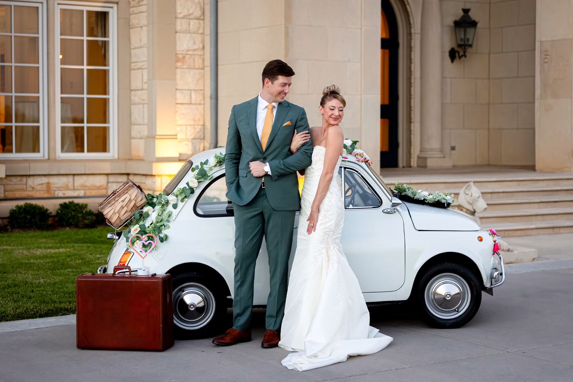 Bride and groom in wedding attire stand beside a decorated white car in front of a building entrance, with a suitcase nearby.