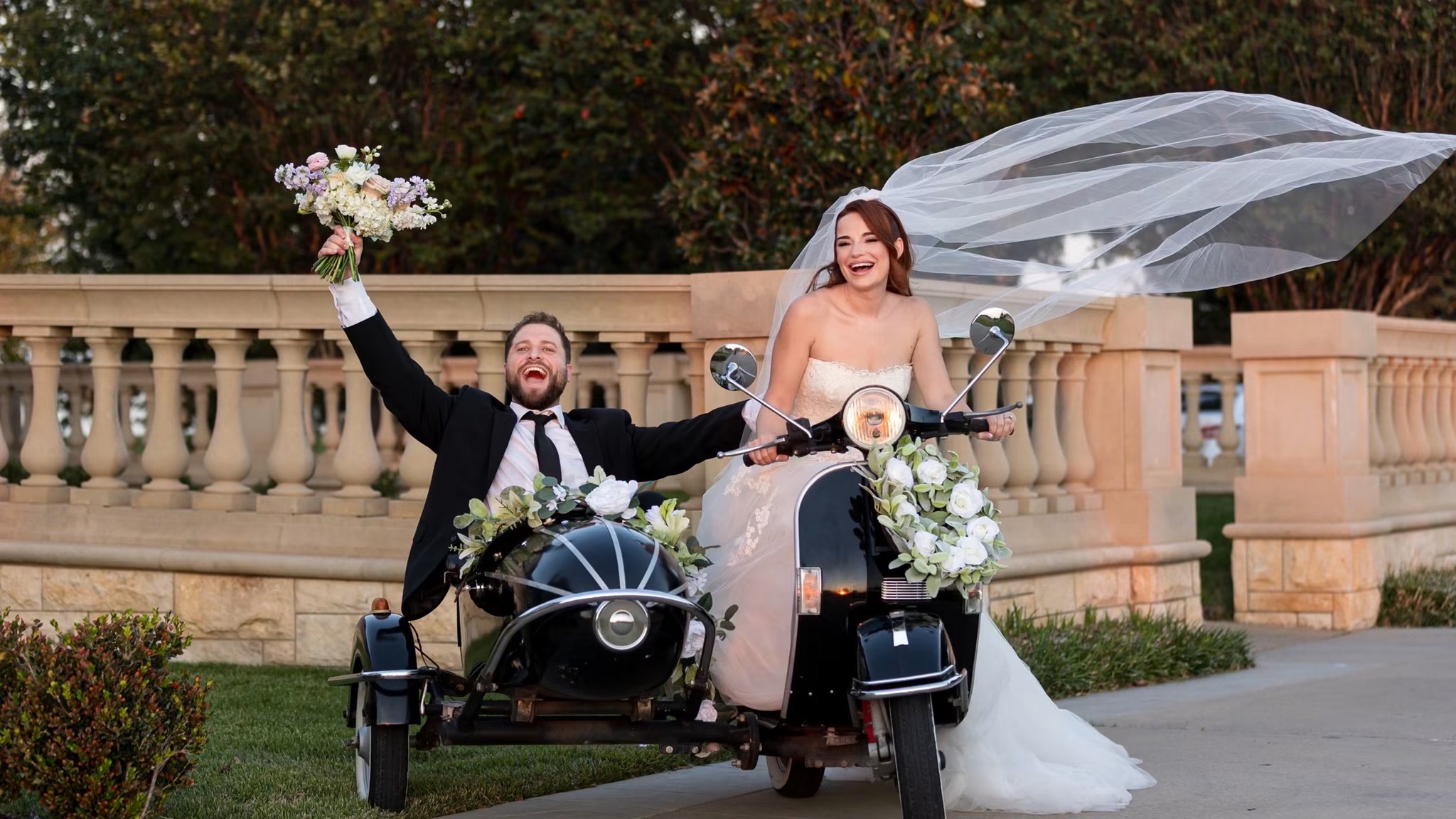 A newlywed couple riding a motorcycle with a sidecar, the bride holding an umbrella and bouquet, outdoors near a stone fence.
