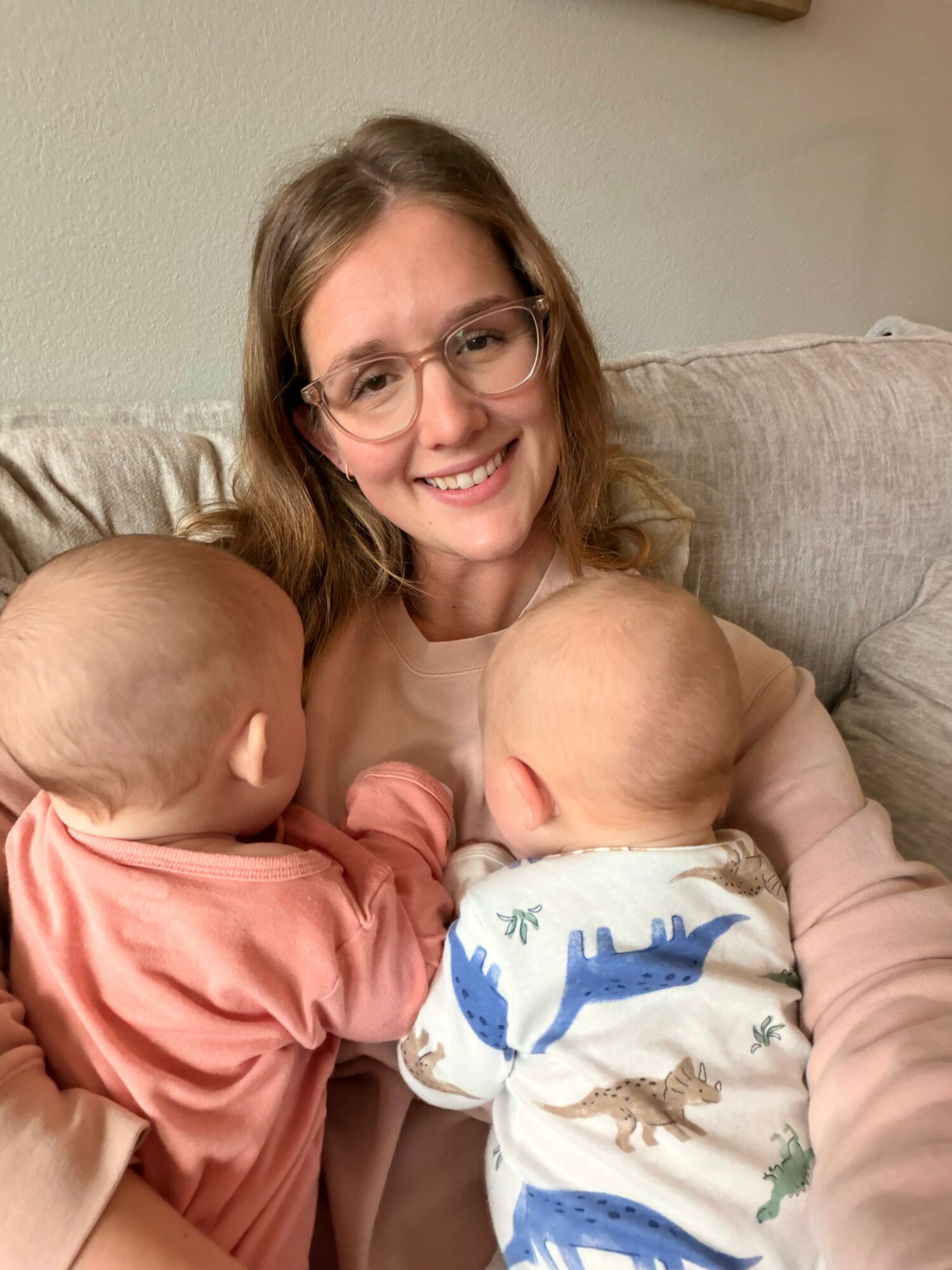Woman with glasses smiling holding two babies, one in each arm, on a beige couch.