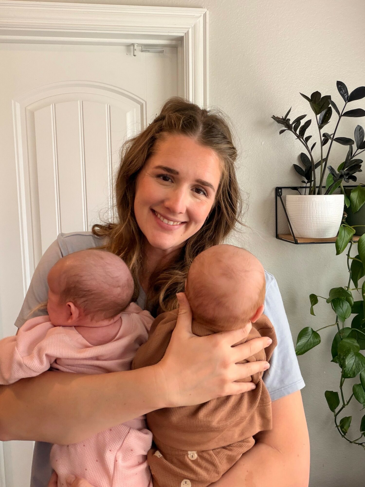 Young woman smiling while holding two babies, one in each arm, indoors near a white door and green plants.