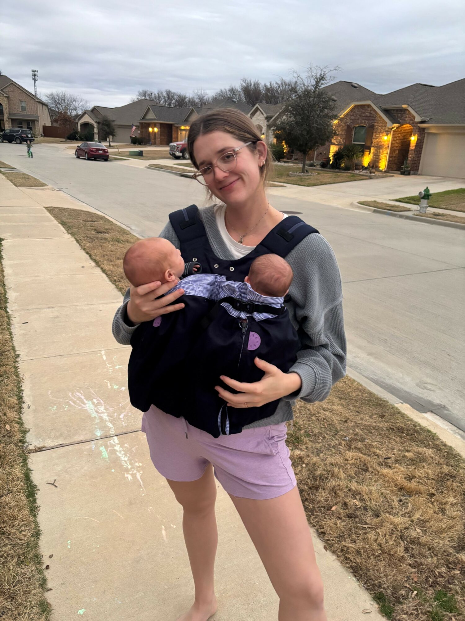 Woman carrying a baby and a doll on a sidewalk, with houses and trees in the background.