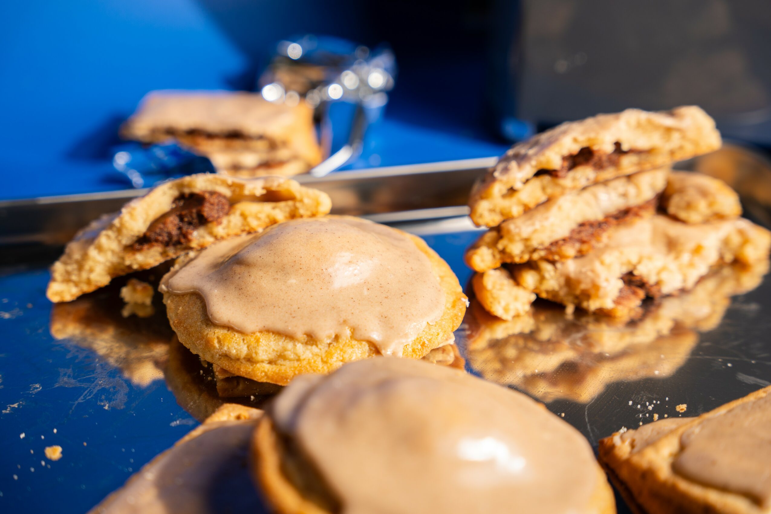 Close-up of cookies with icing on a reflective surface, some broken revealing filling inside, with a blurred background.