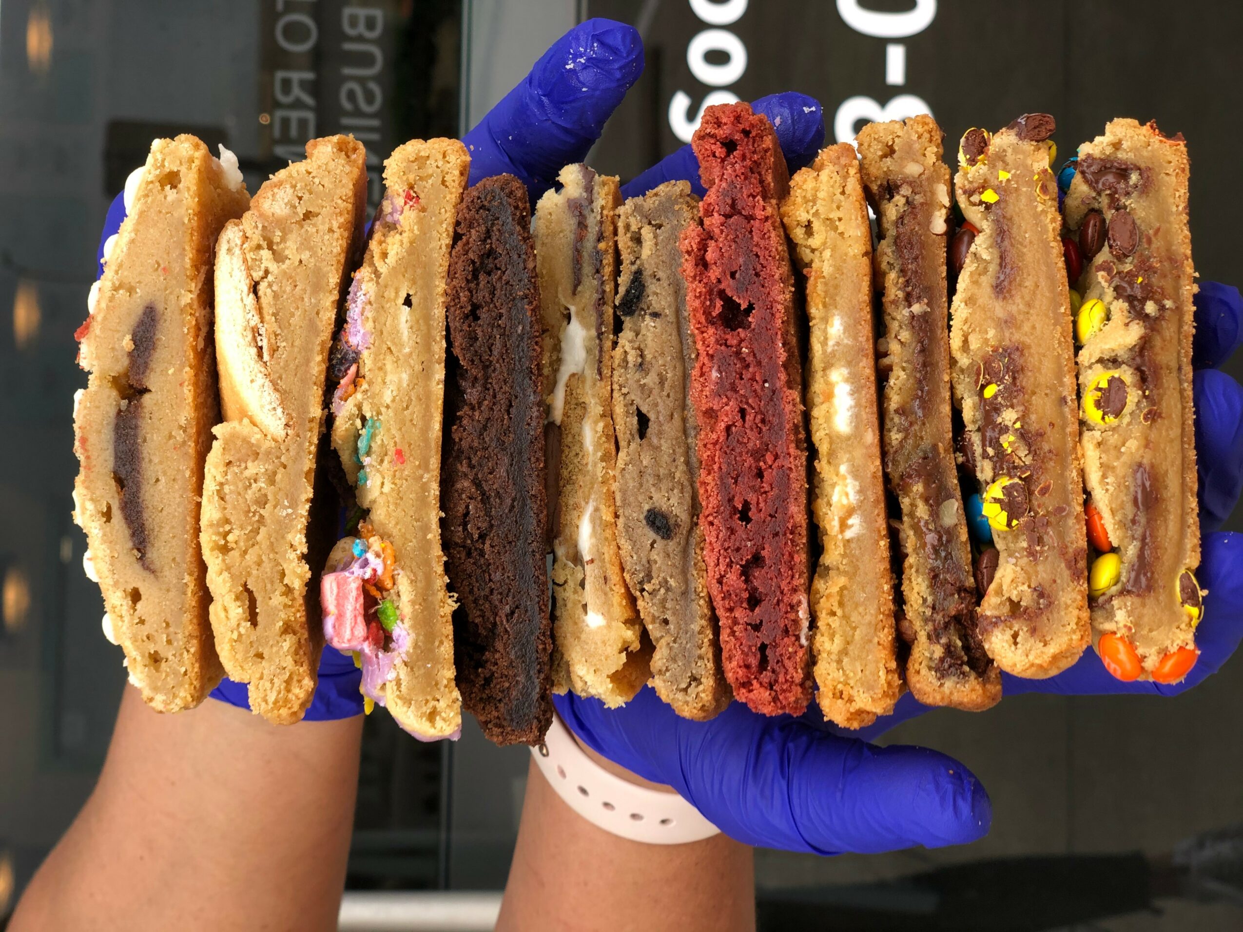 Person wearing a blue glove holding a row of cookies with colorful candies and chocolate chips, background of a store.