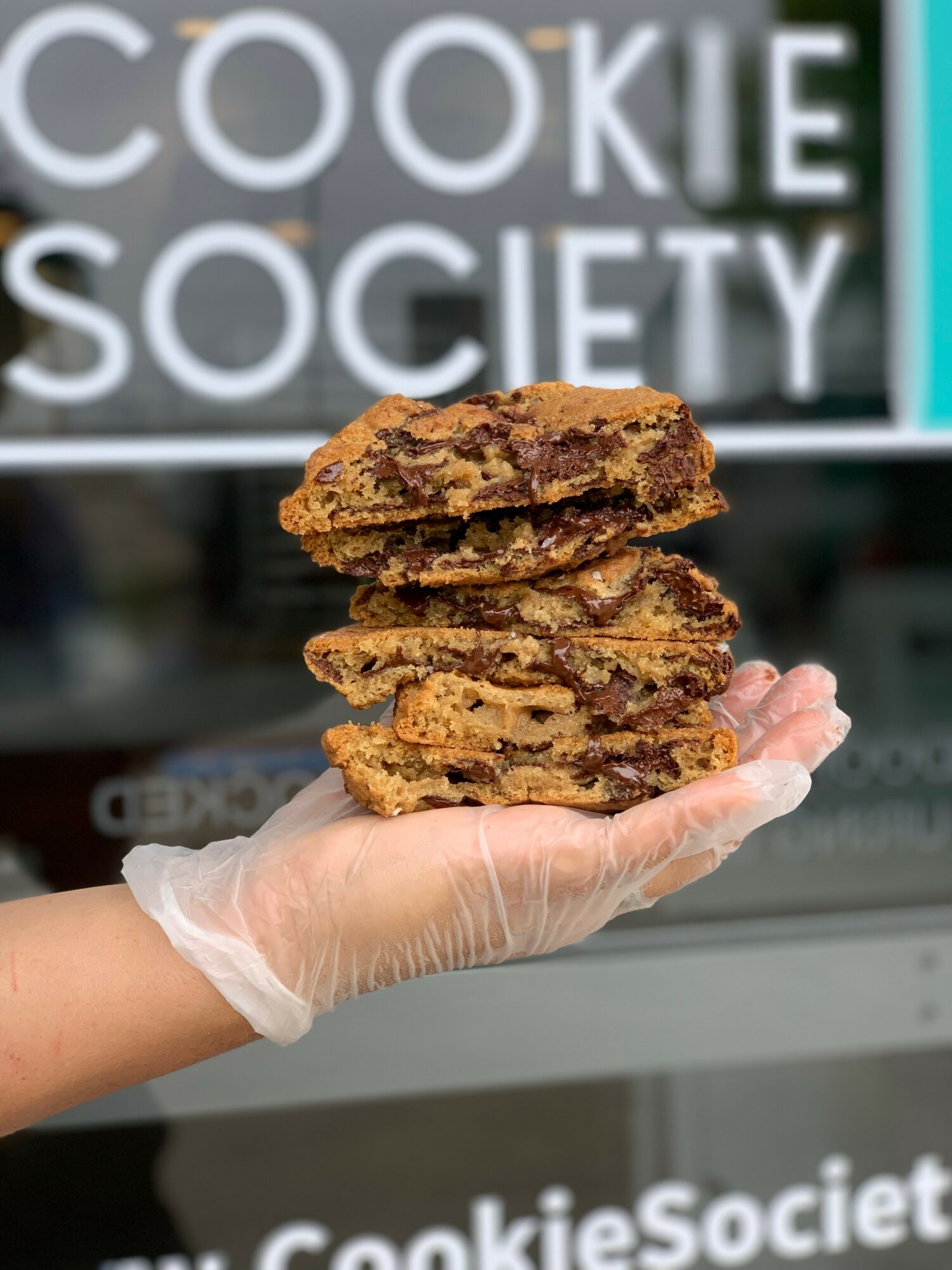 Hand in a glove holding four stacked cookies in front of a sign that reads Cookie Society.