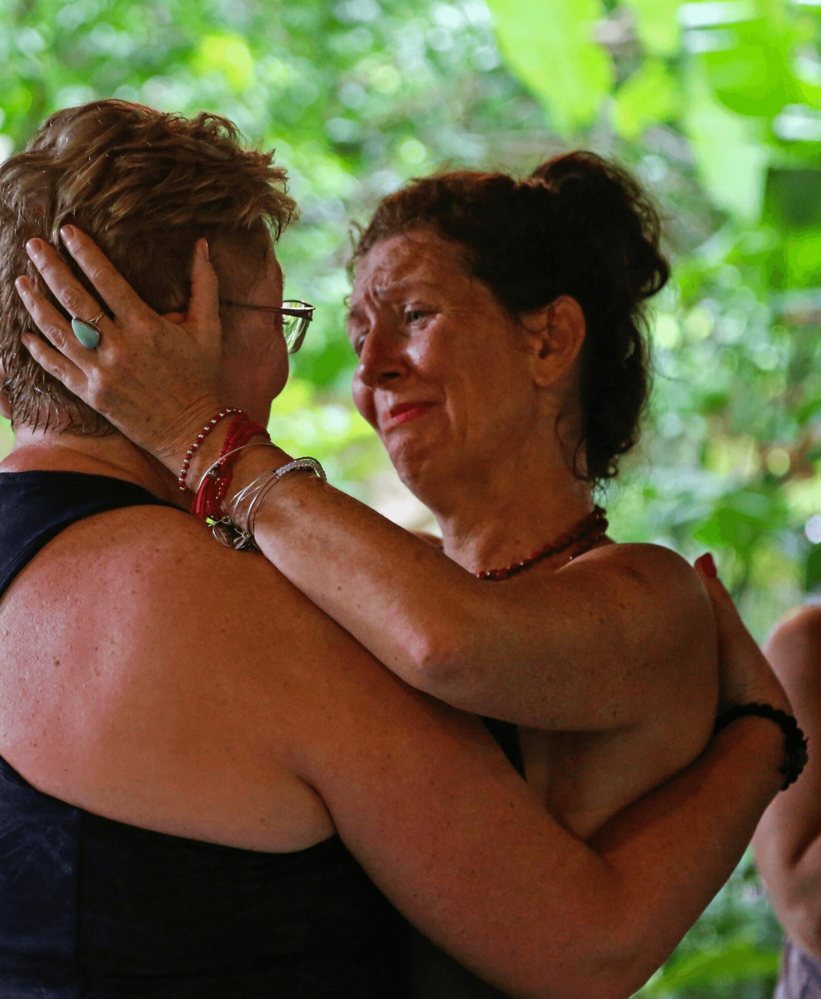 Two women embrace, one with glasses and short hair, the other with curly hair, smiling in a green outdoor setting.