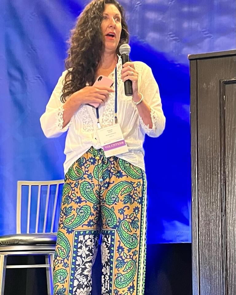 Woman with curly hair speaking into a microphone, standing in front of a blue backdrop, wearing a white top and colorful patterned pants.