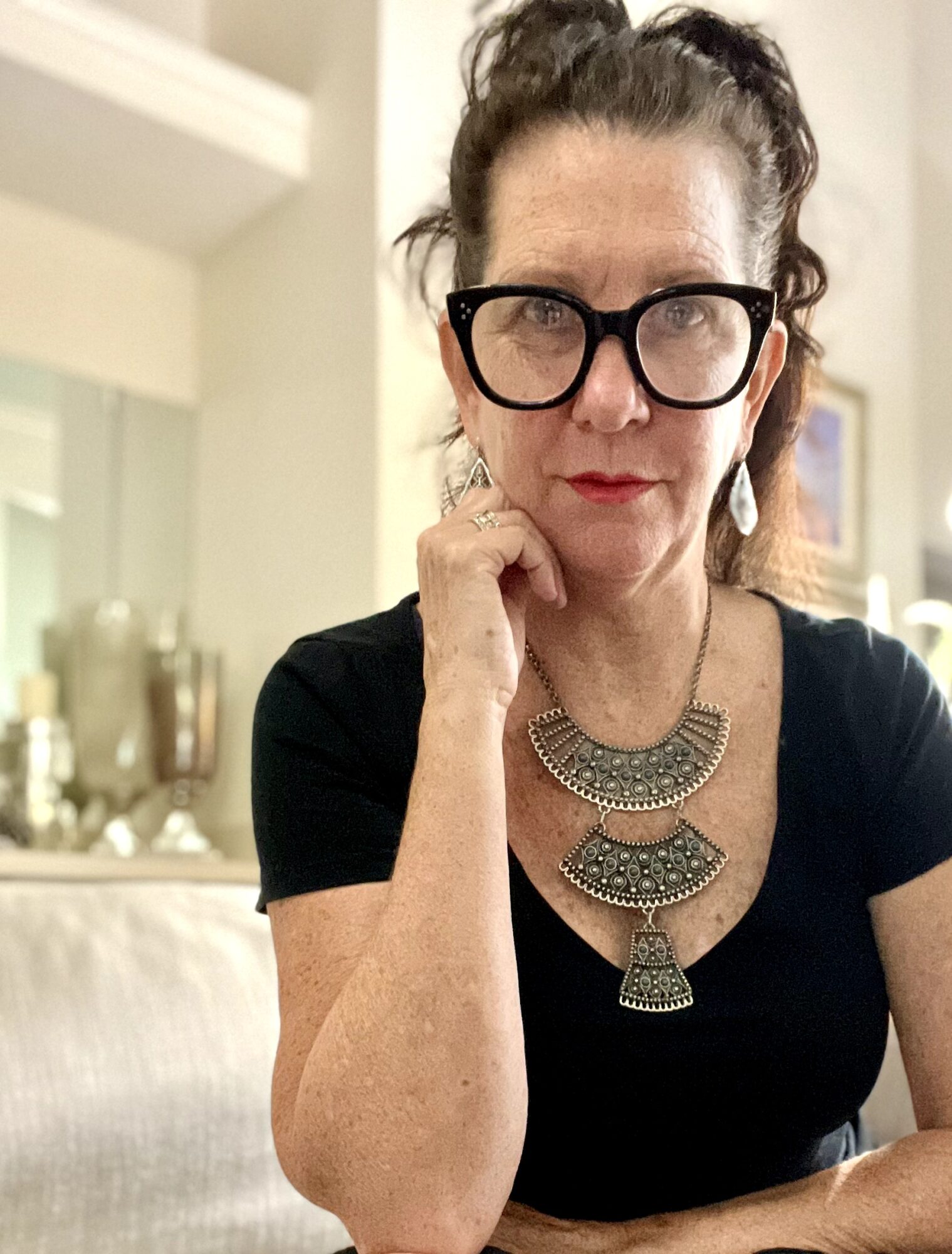 Woman with glasses and dark hair in a bun, wearing a black top and statement necklace, resting her chin on her hand.