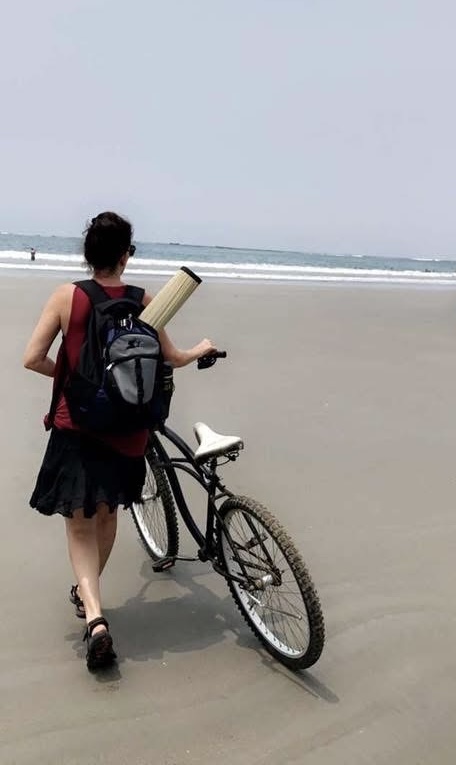 Person walking on beach with bicycle, carrying a book, facing away from camera, ocean in background.