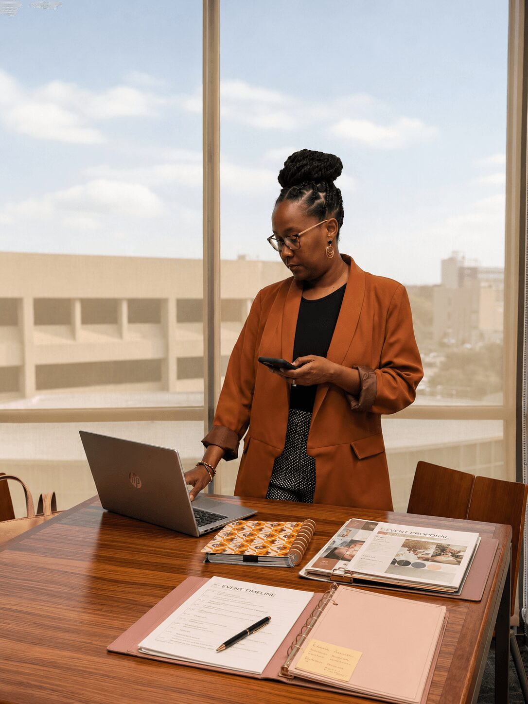 Woman with glasses and braided hair in a brown blazer looks at her phone in a conference room with a laptop and documents on the table.