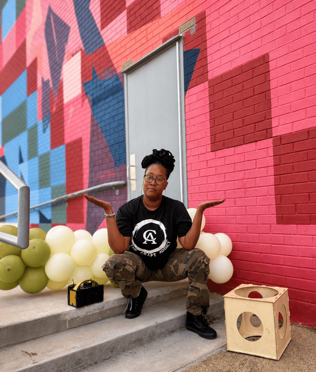 Person sitting on steps with arms raised, colorful mural wall behind, balloons, and a wooden box nearby.