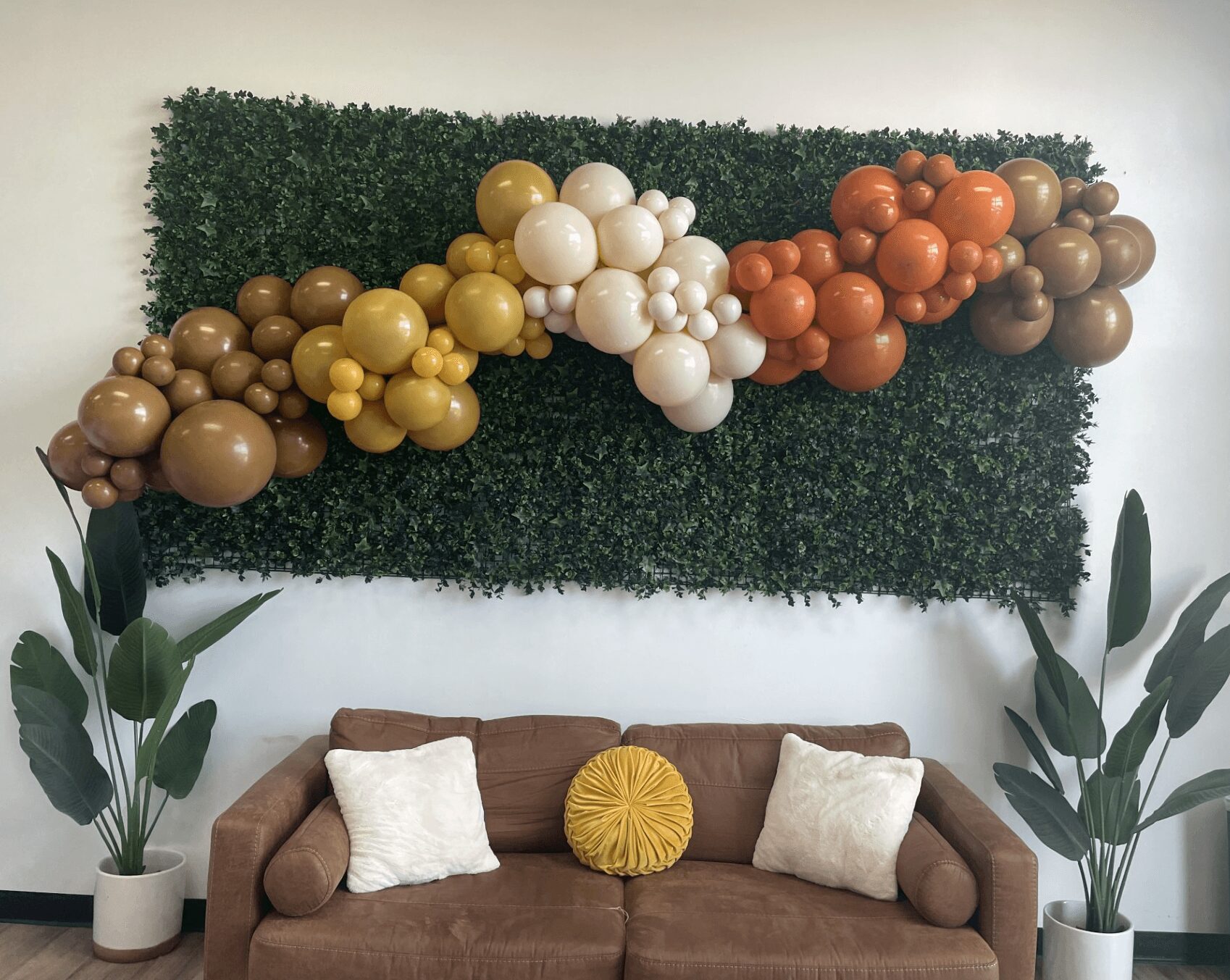 Decorative balloon arrangement on a green hedge wall above a sofa with pillows, flanked by potted plants.