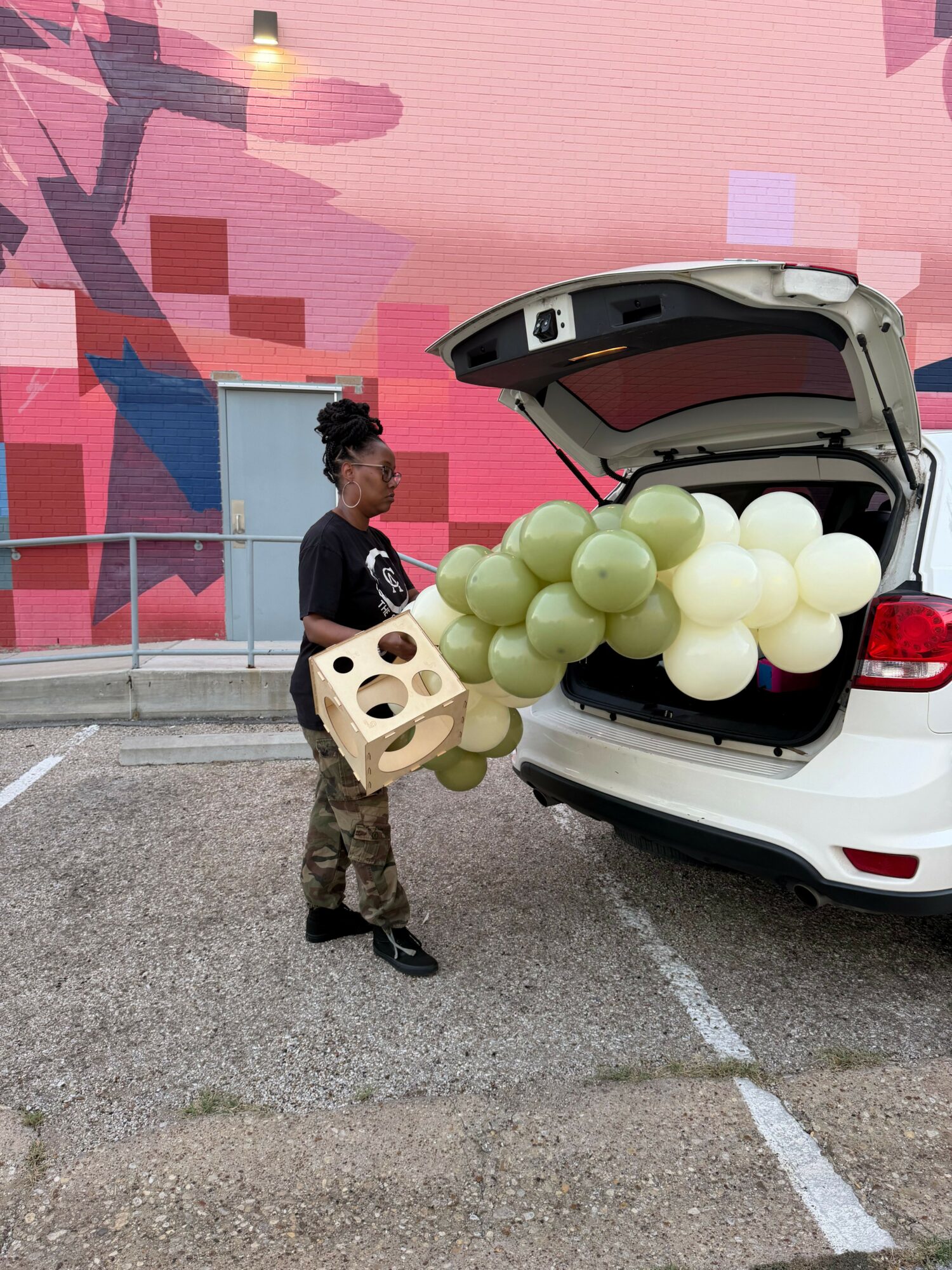 Person loading balloons into the back of a white car in front of a pink wall.
