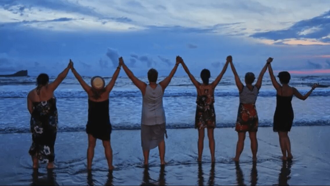 Six people holding hands on the beach at sunset, facing the ocean, with waves in the background.