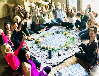 Group of people sitting in a circle on the floor, celebrating around a decorated circular cloth, with some raising their hands.