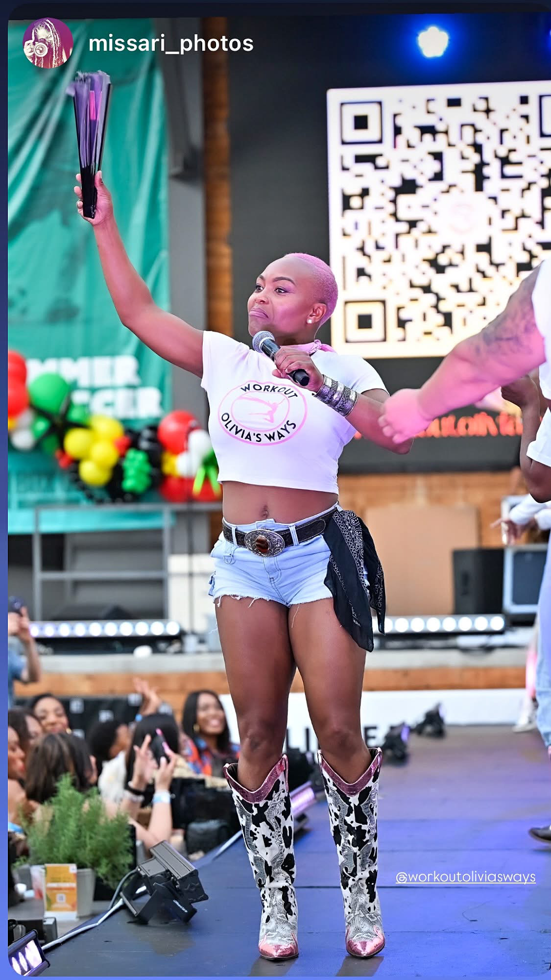 Young woman on stage holding a trophy, wearing a white crop top, denim shorts, and patterned boots, with a large QR code behind her.