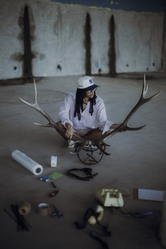 Person with long hair and helmet sitting on the ground holding large antlers, surrounded by tools and objects.