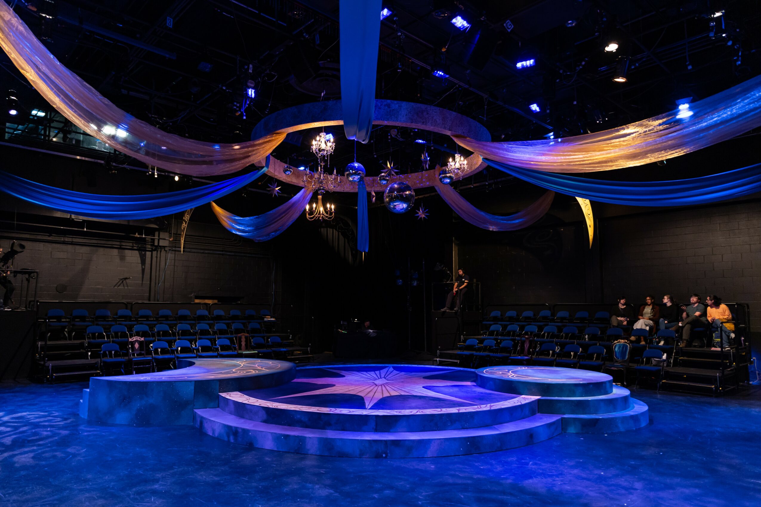 Stage with a star design, draped fabric, and chandeliers in a dark, empty auditorium with seating on both sides.