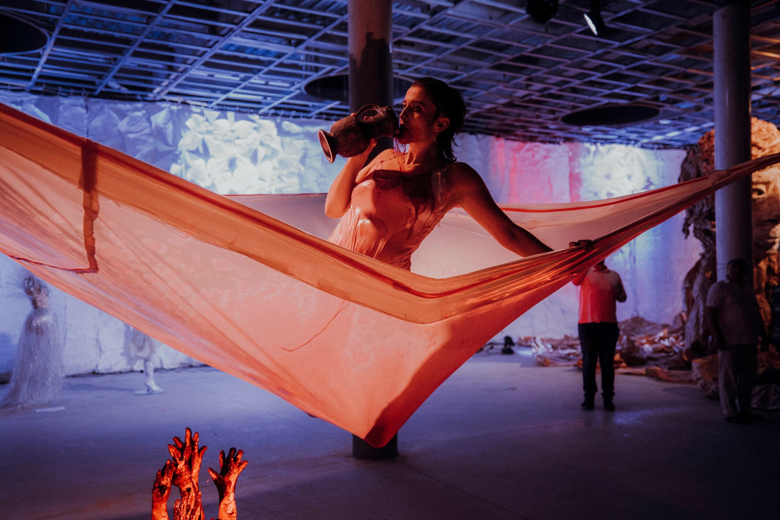 Person lying on a large fabric hammock, holding a drink, in an indoor space with colorful lighting and other people present.