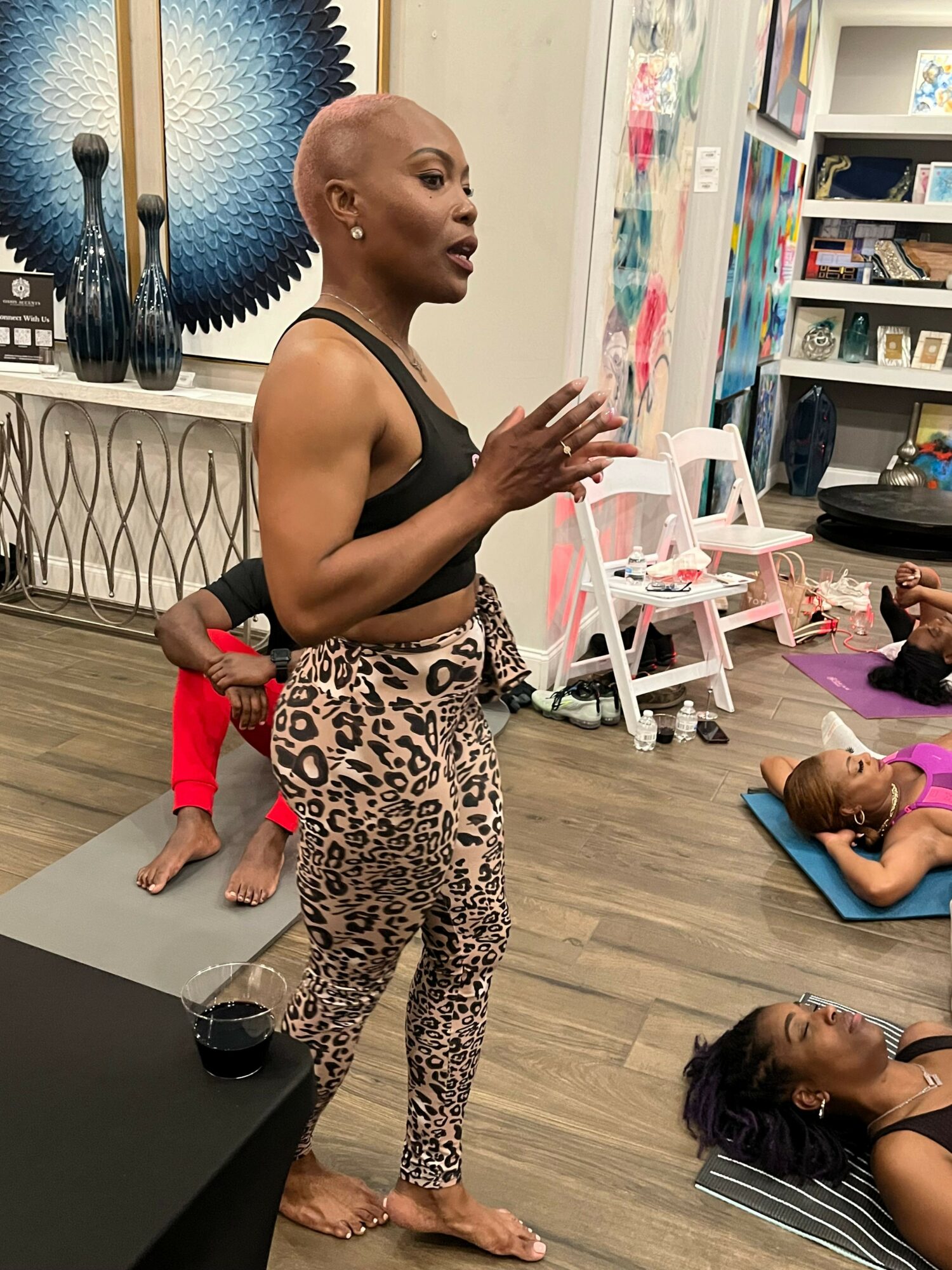 Group of people practicing yoga or stretching on a wooden floor in a room with shelves and colorful items, with a child in red nearby.