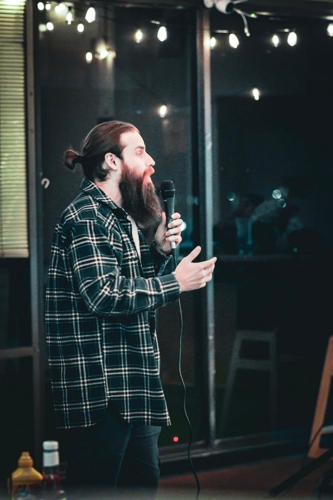 Man with long hair and beard holding a microphone, standing indoors near a window with lights outside.