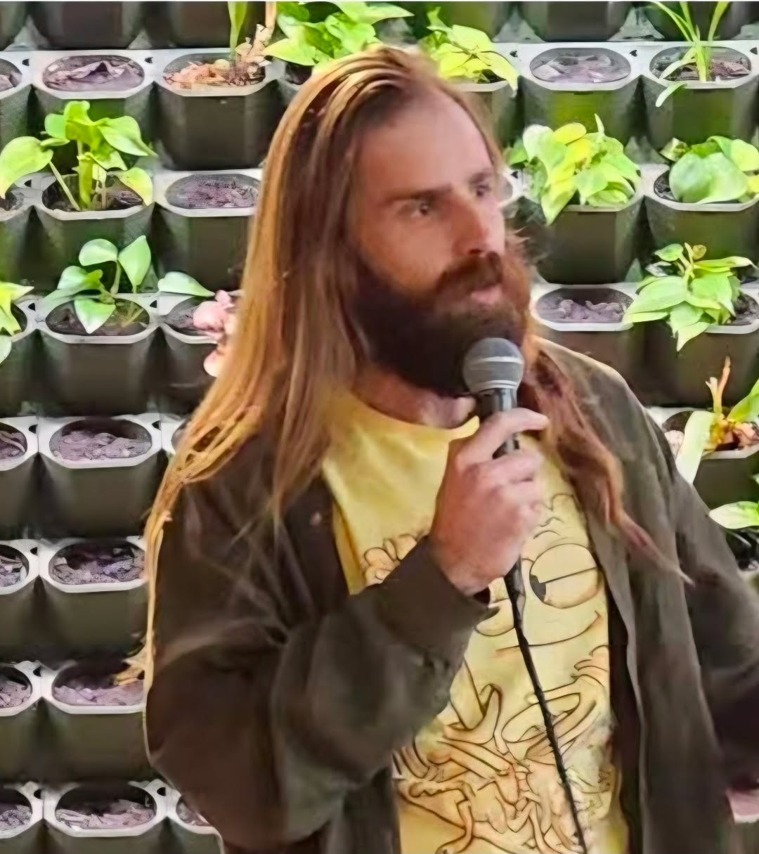 Man with long hair and beard holding a microphone, standing in front of a wall of potted plants.