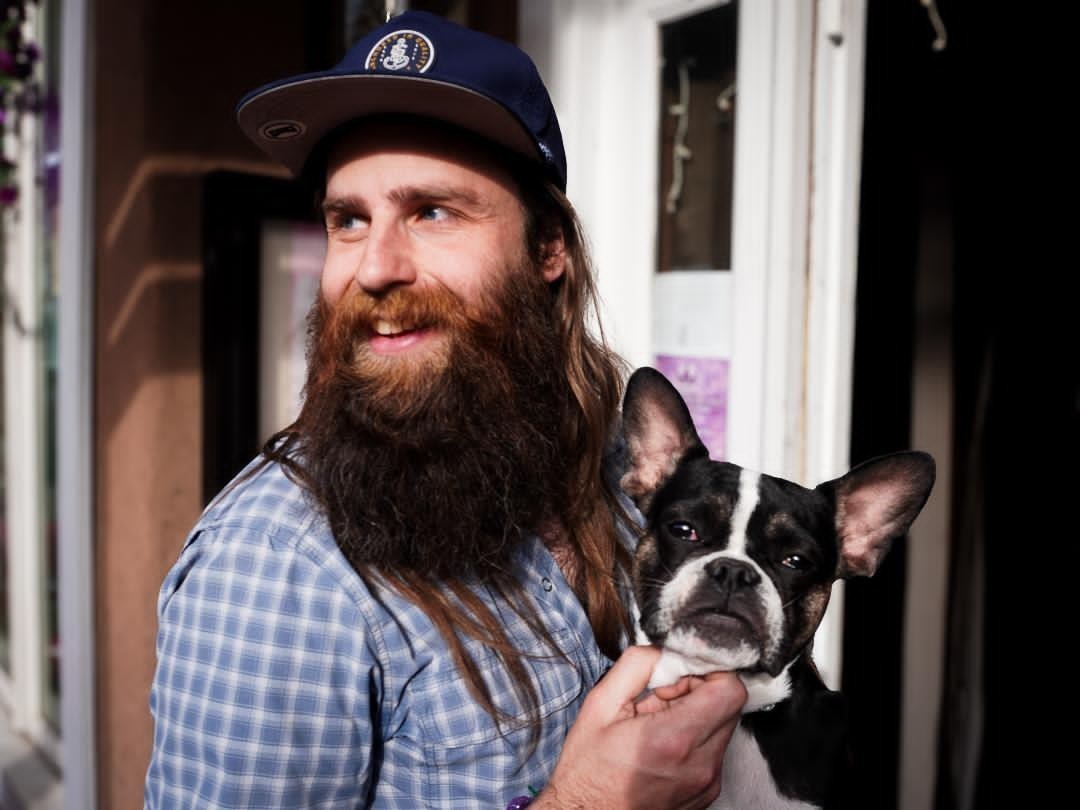 Man with long hair and beard holding a black and white dog, smiling, in front of a window.