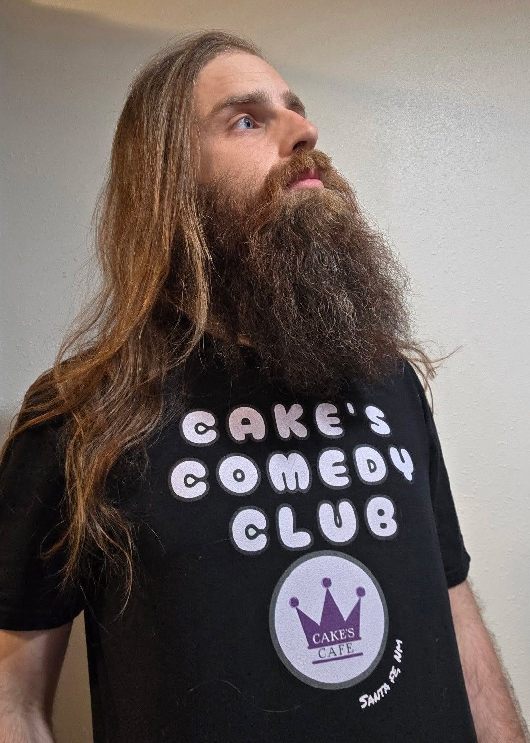 Man with long hair and beard wearing a black T-shirt with white text and a purple crown logo.