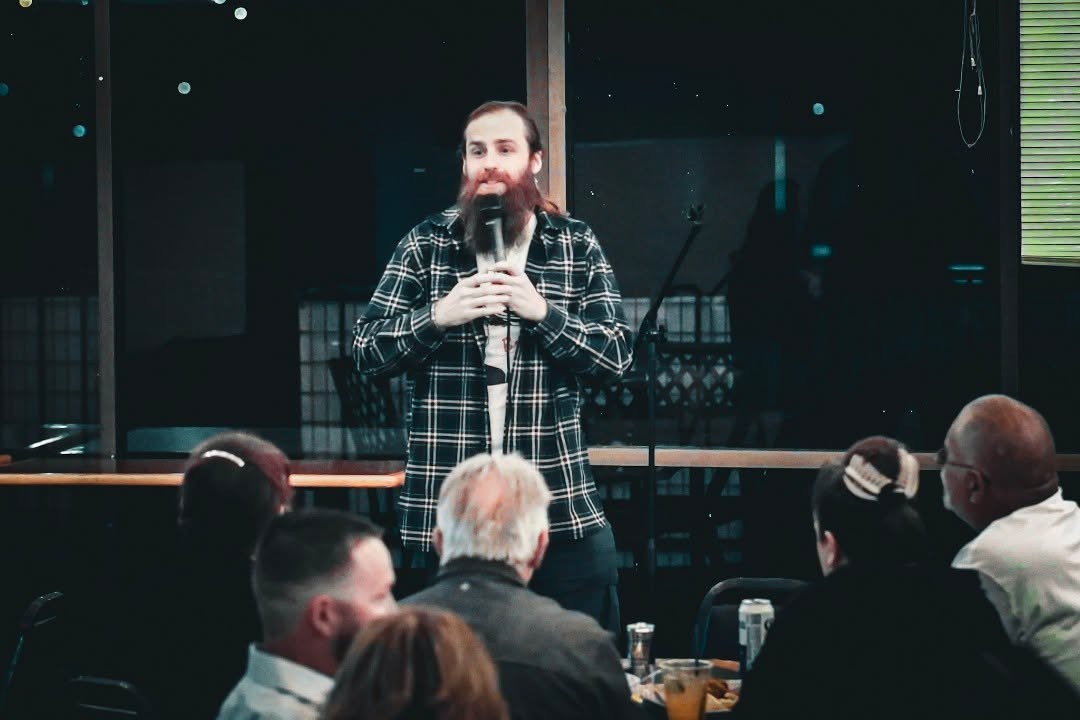 A man with a beard and plaid shirt stands on stage holding a microphone, speaking to an audience in a dimly lit room.