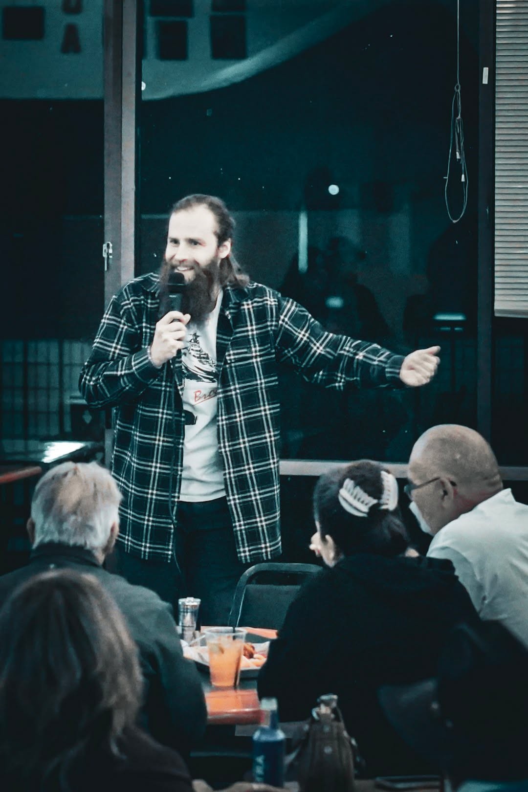 Man with a beard and plaid shirt speaking into a microphone in front of an audience, gesturing with his right hand.