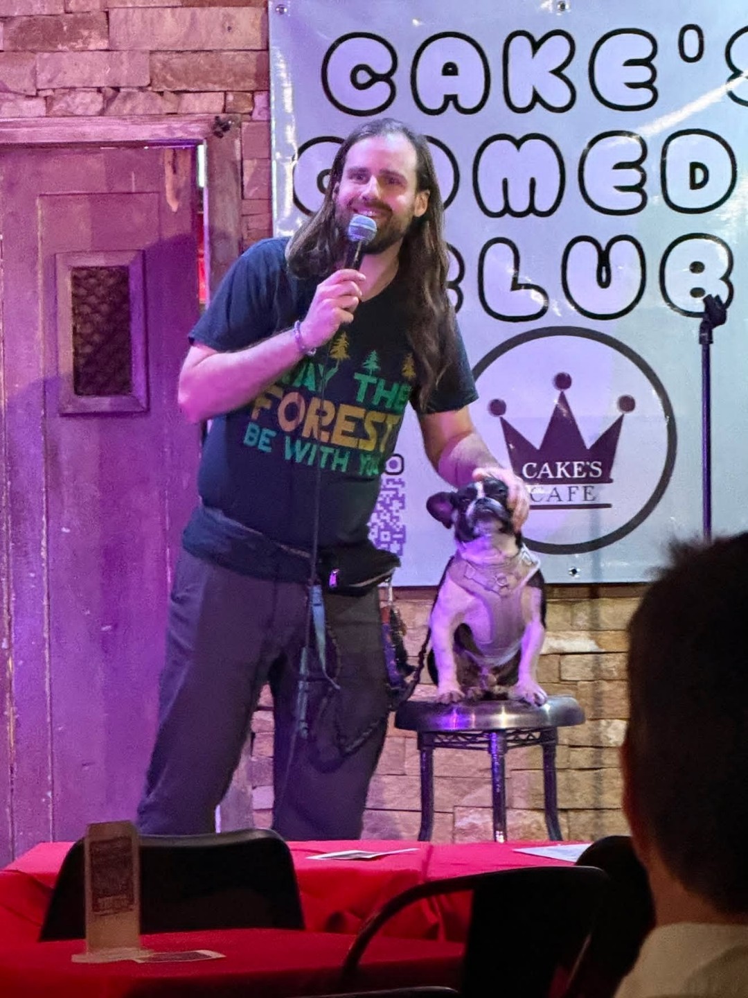 Man with long hair and beard holding a microphone on stage with a dog sitting on a table, background with signs and brick wall.