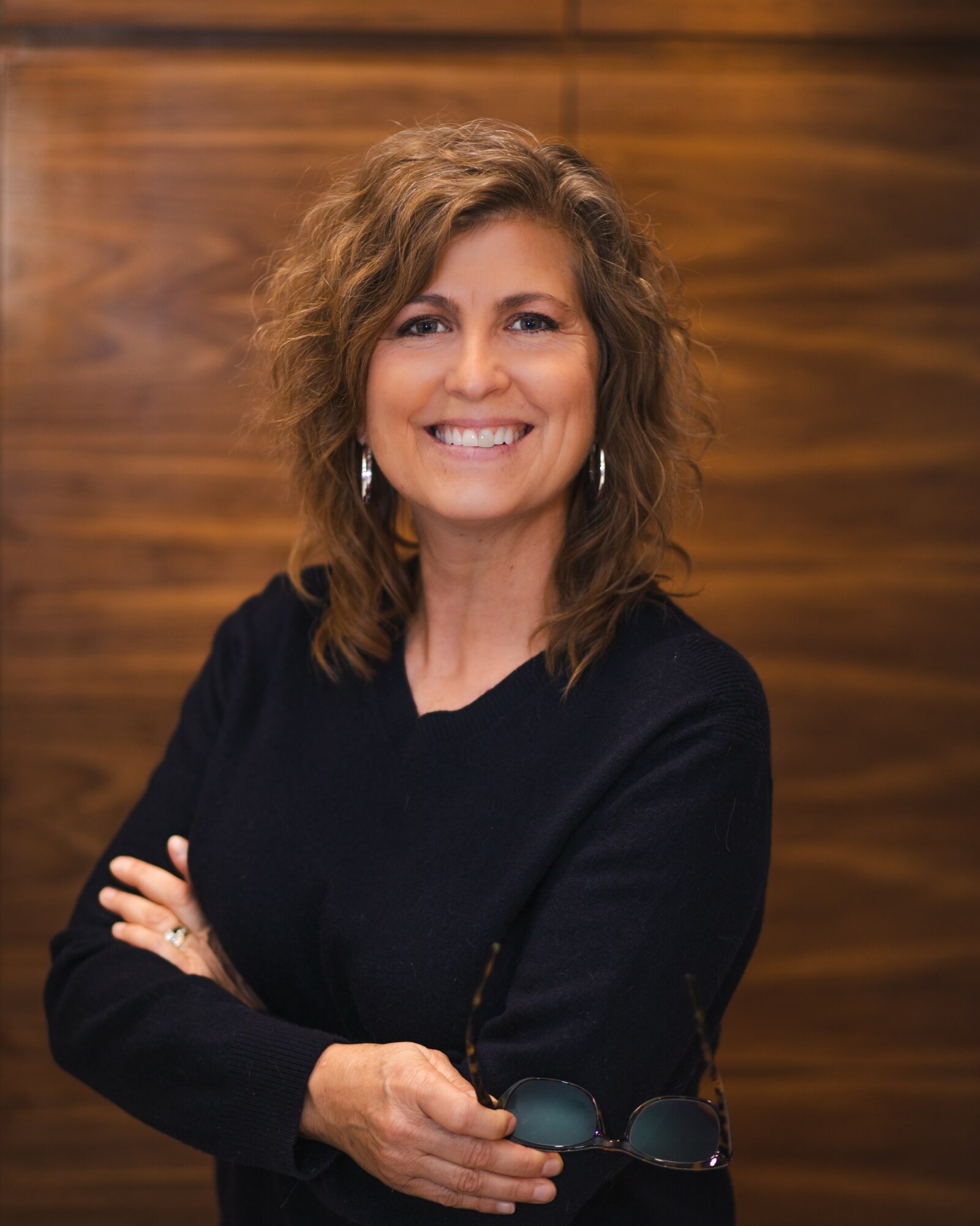 Woman with curly hair smiling, wearing a black top, standing against a wooden background.