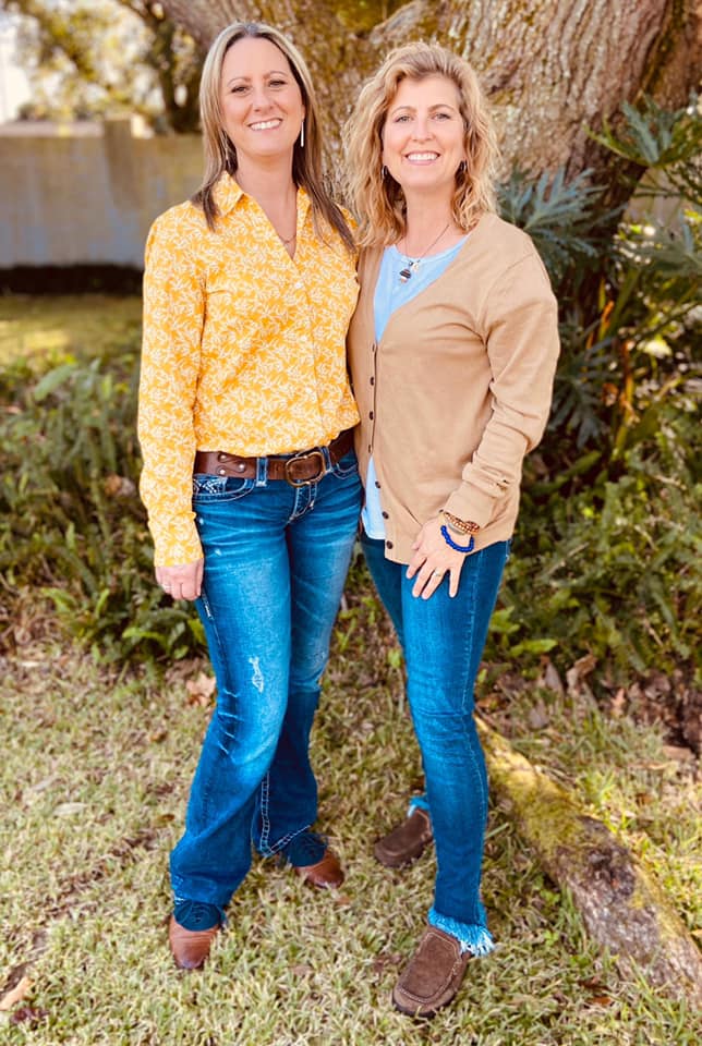Two women standing outdoors, smiling, with trees and plants in the background.