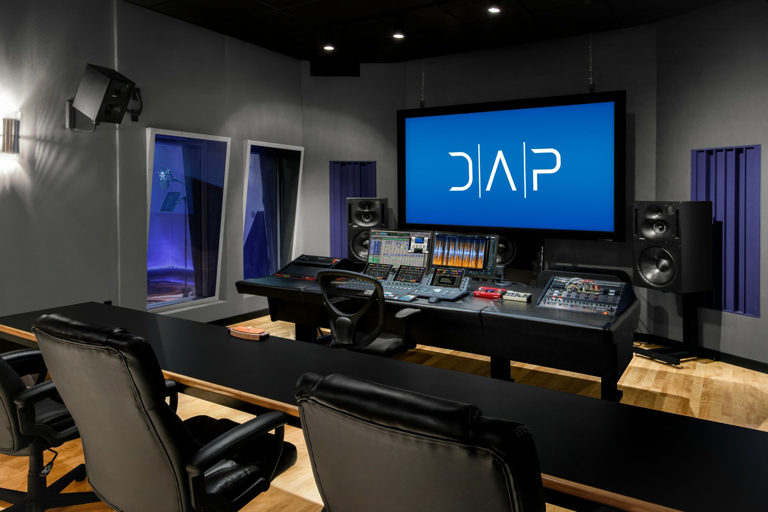 Recording studio with mixing console, large screen displaying 'DAP', and sound equipment, chairs in foreground, windows on side walls.