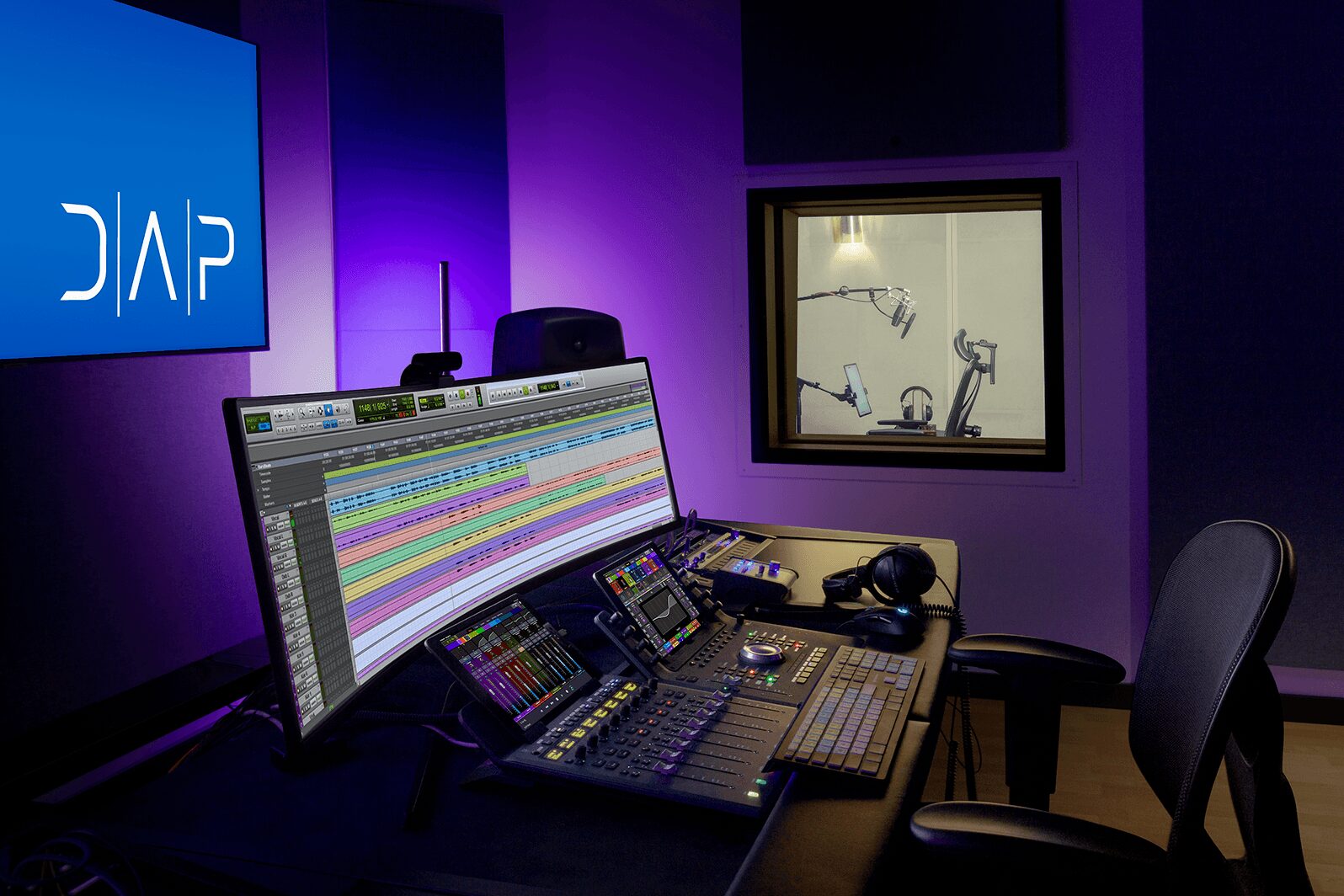 Recording studio with mixing console, computer monitor, and window showing a microphone and headphones, illuminated by purple lighting.