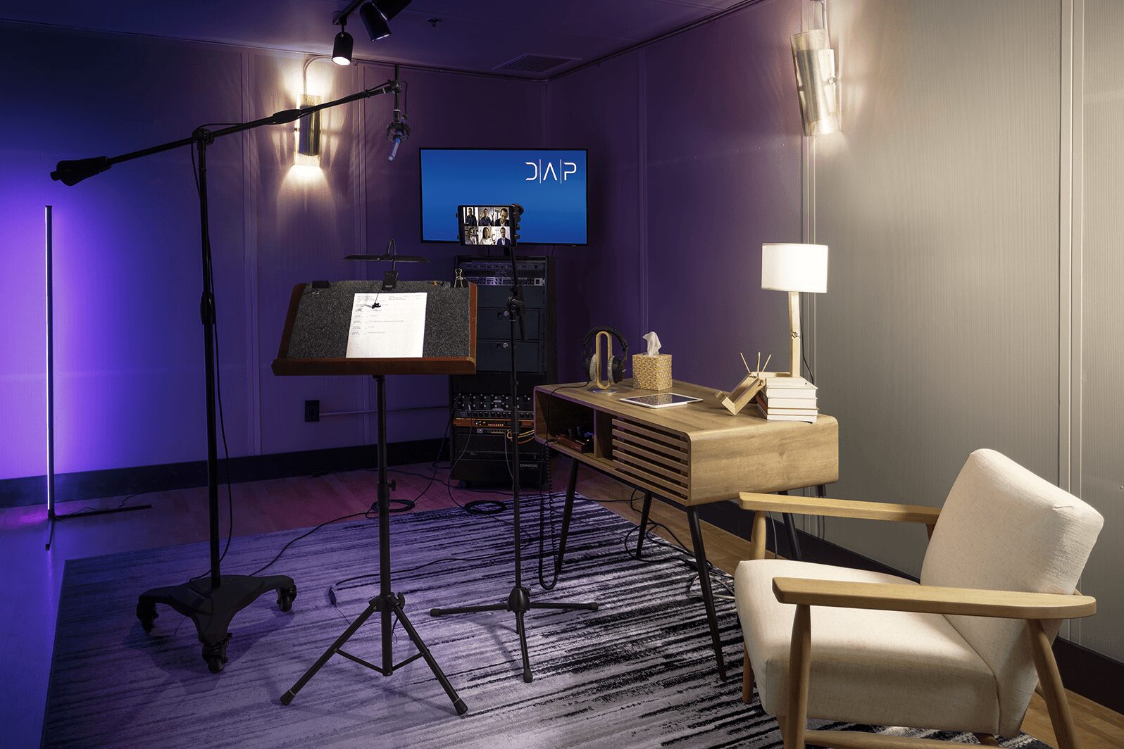 Recording studio with microphone, music stand, TV screen, table lamp, and armchair, illuminated by wall lights.