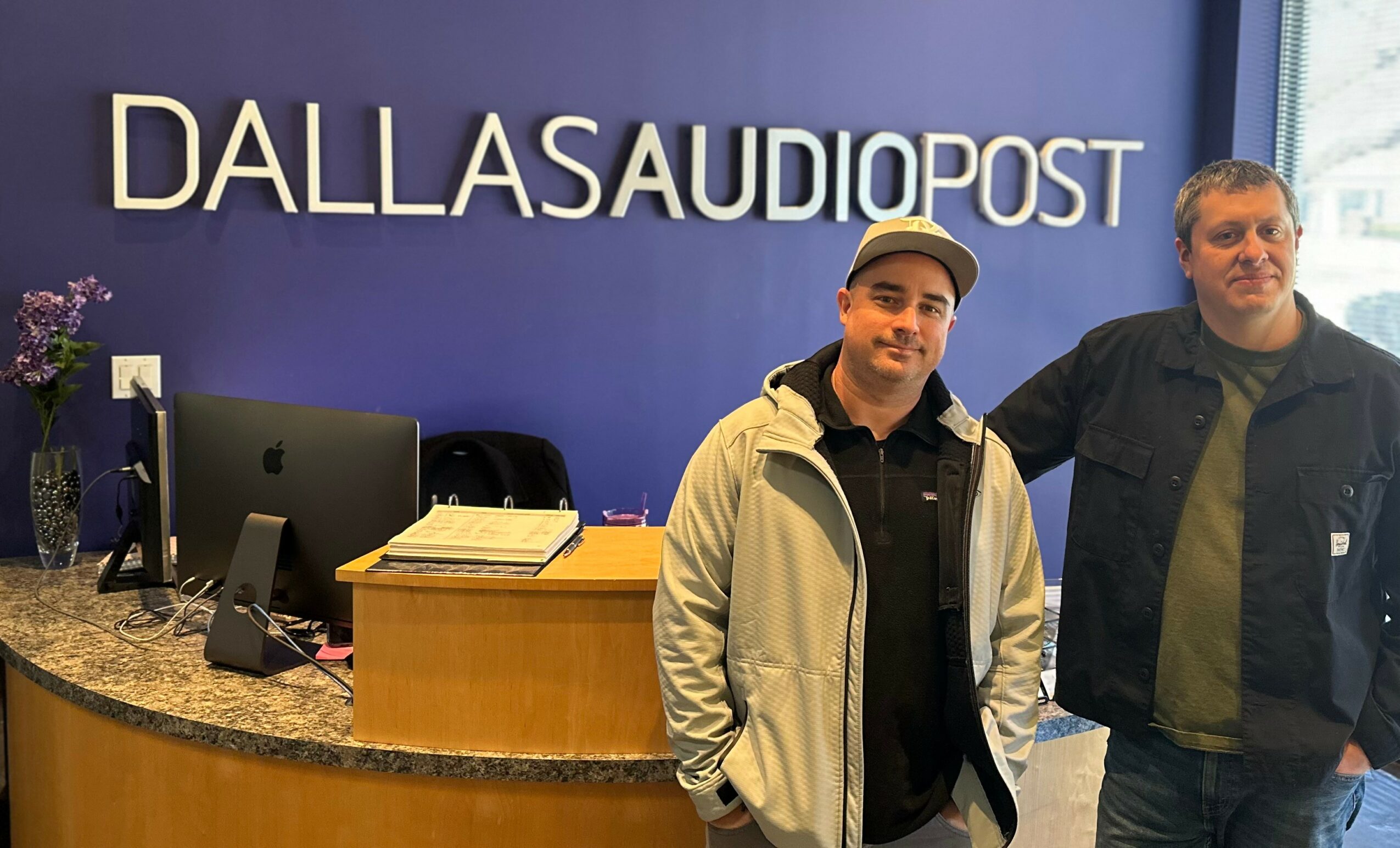 Two men standing in front of a reception desk at Dallas Audio Post, with a blue wall and sign behind them.