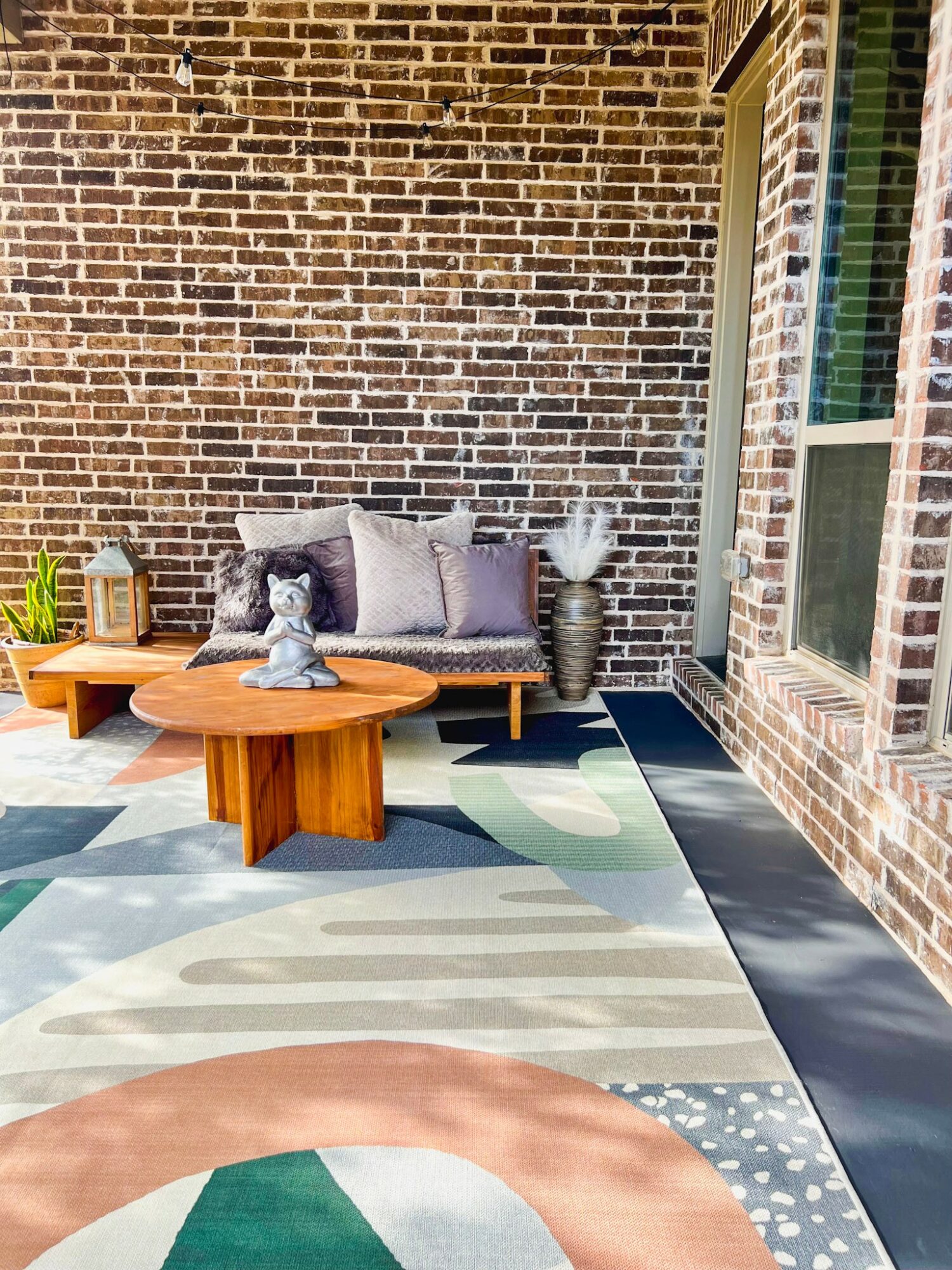 Outdoor patio with brick wall, sofa with cushions, wooden coffee table, and patterned rug, sunlight streaming through window.