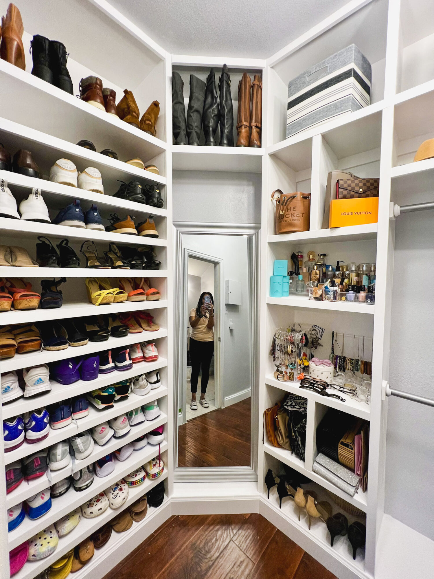 Closet with shelves filled with shoes, bags, and accessories, and a full-length mirror reflecting a person taking a photo.