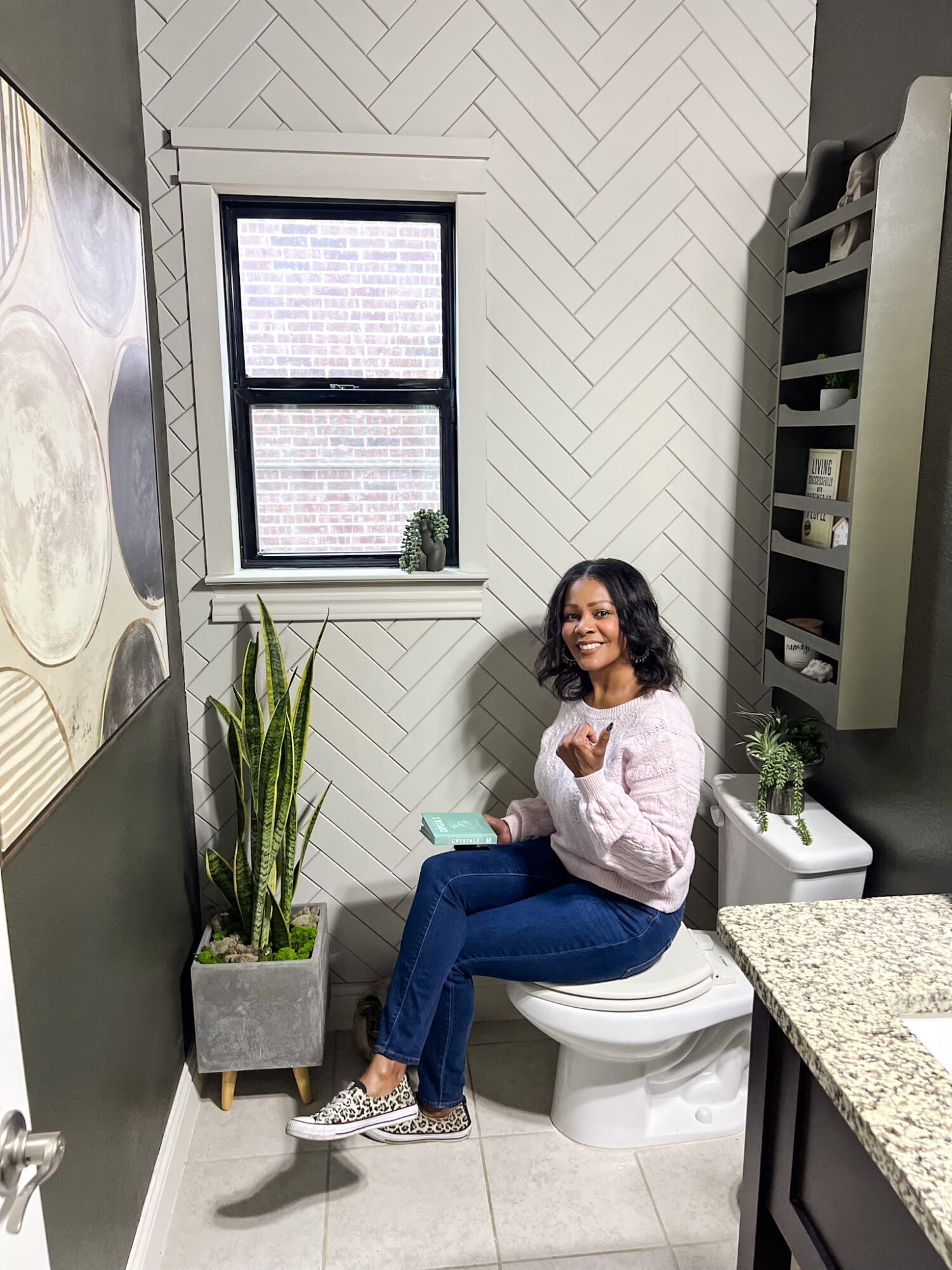 Woman sitting on toilet in bathroom, smiling, holding a phone, with plants and window behind her.