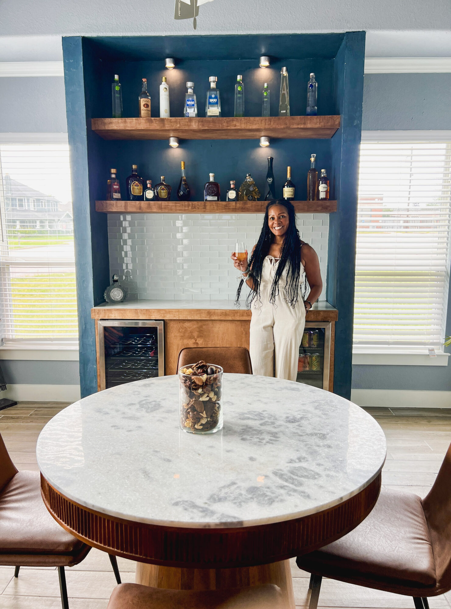 Woman in white dress standing behind a marble-topped table in a room with a blue wall and shelves with bottles.