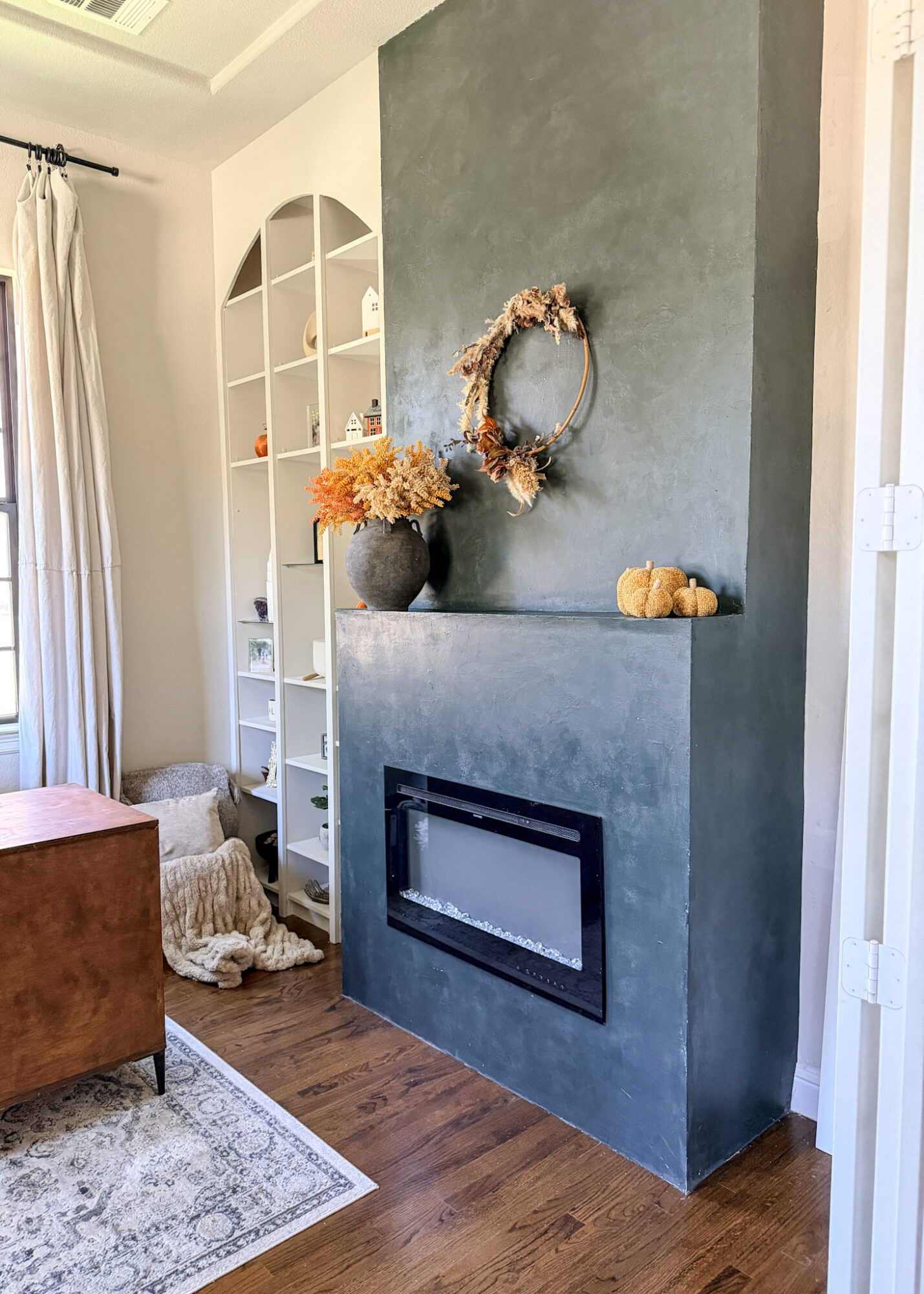 Interior corner with a blue wall, fireplace, decorative wreath, vase with dried flowers, and a white bookshelf, with a window and curtain nearby.