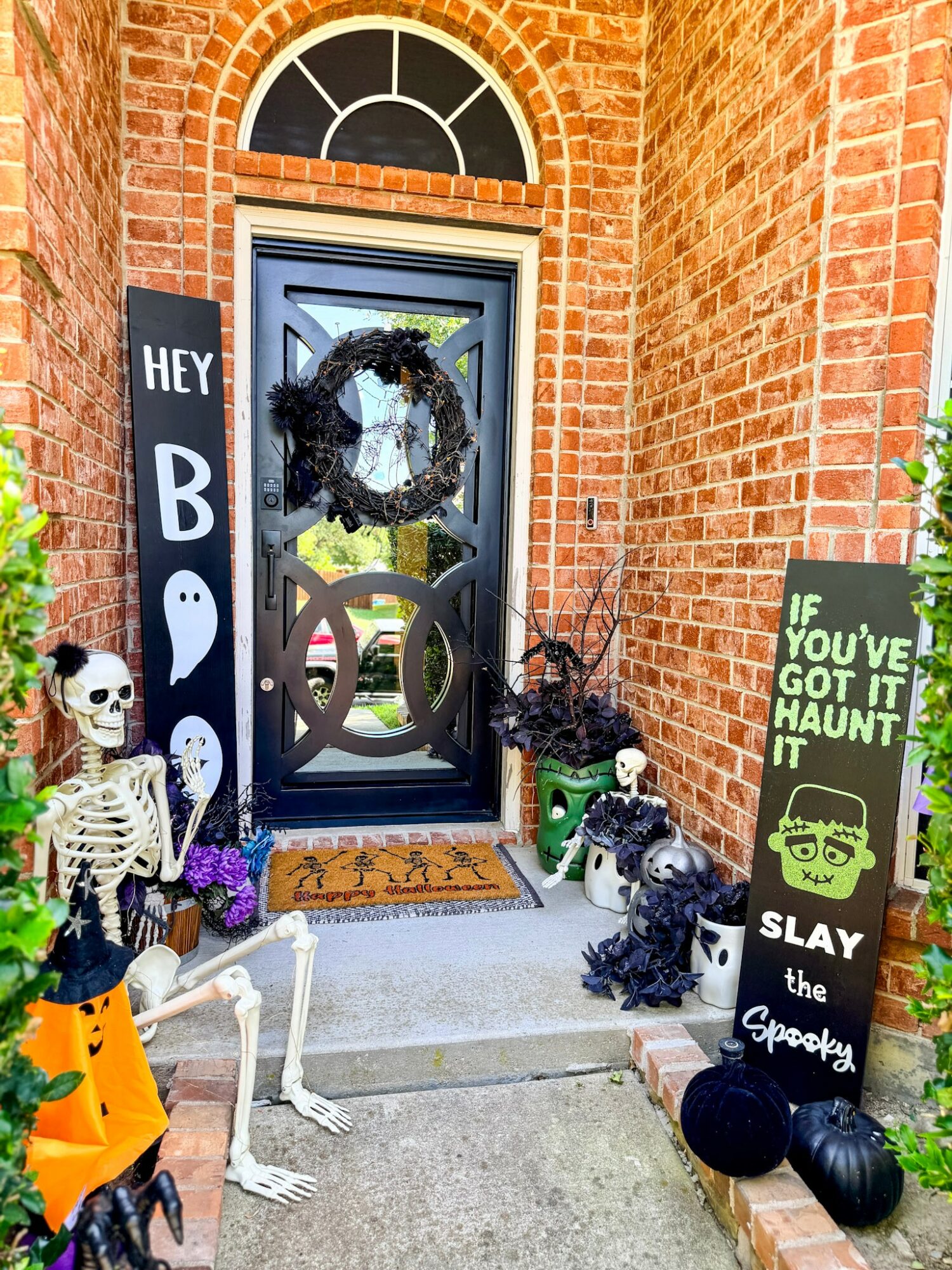 Front door decorated with Halloween themed skeletons, pumpkins, and signs, with a wreath on the door and a welcome mat.