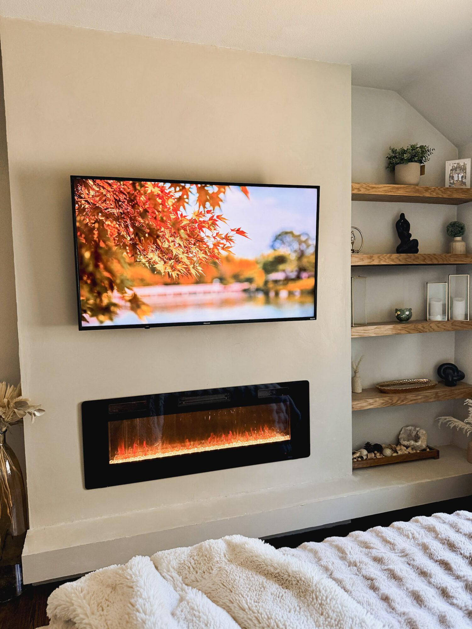 Living room with wall-mounted TV displaying autumn leaves and a modern electric fireplace below it, with shelves on the right side.