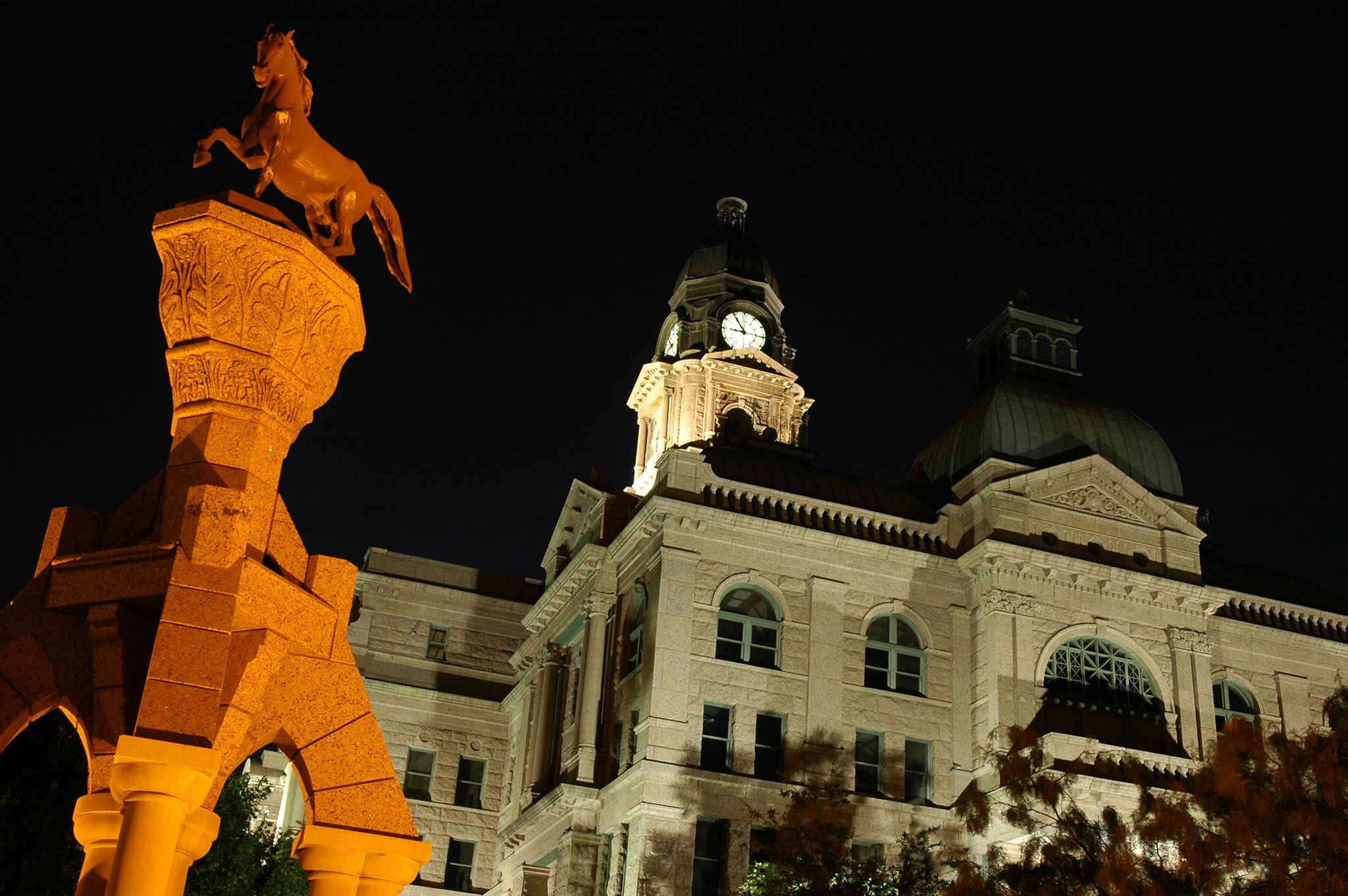 Nighttime view of a historic building with a clock tower and a statue illuminated in orange light, dark sky background.