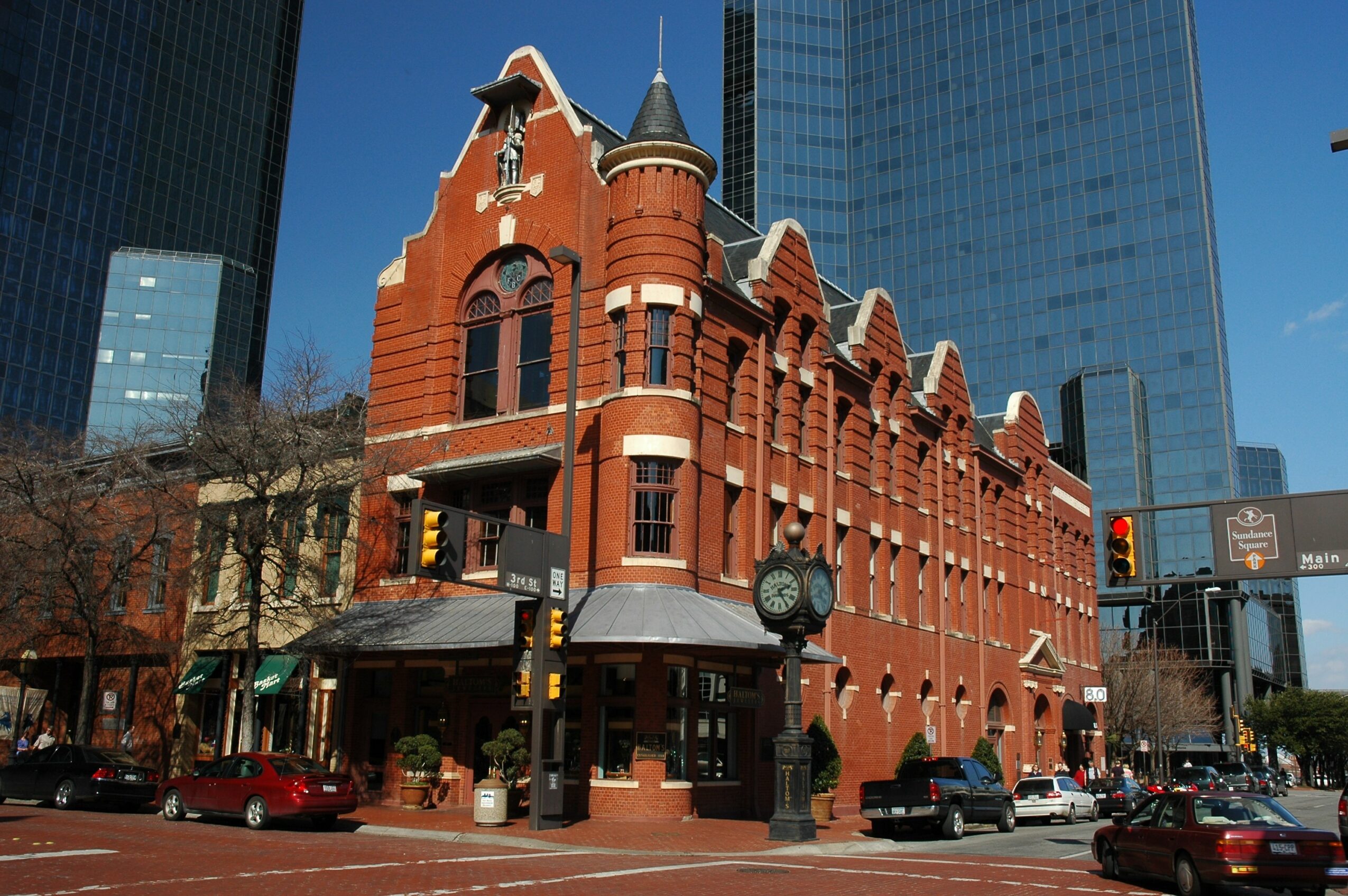 Red brick historic building with turrets and arched windows, surrounded by modern glass skyscrapers, on a city street corner.