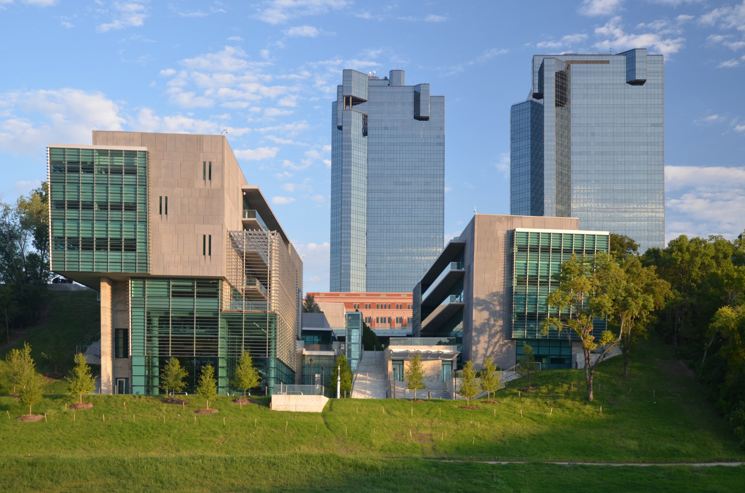 Modern buildings with glass facades and greenery under a blue sky with clouds.