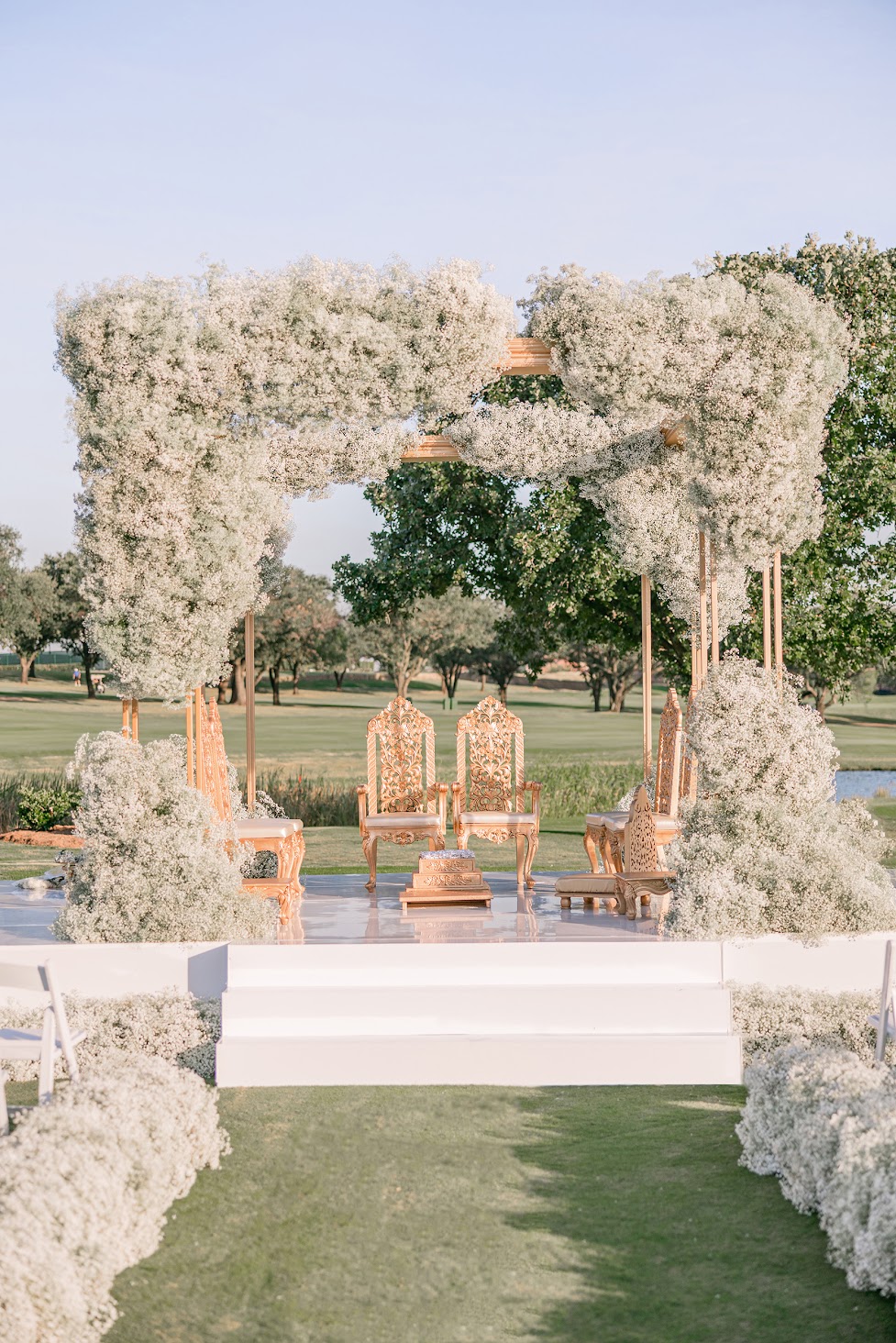 Decorative floral arch with chairs and pathway outdoors, greenery in background, clear sky, wedding setup.